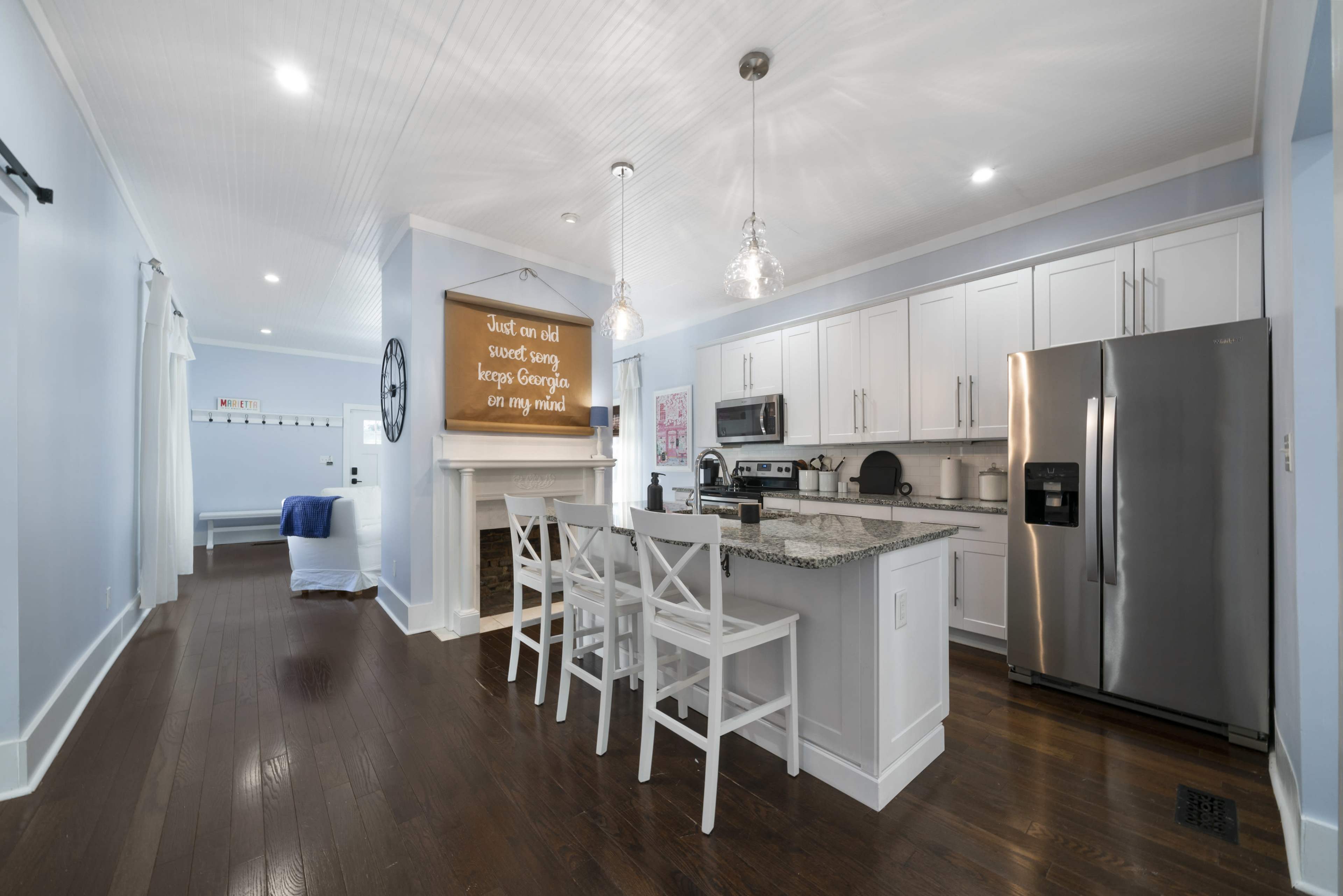 The image shows a modern kitchen featuring white cabinetry, a granite countertop, stainless steel appliances, and a small dining area with white chairs.