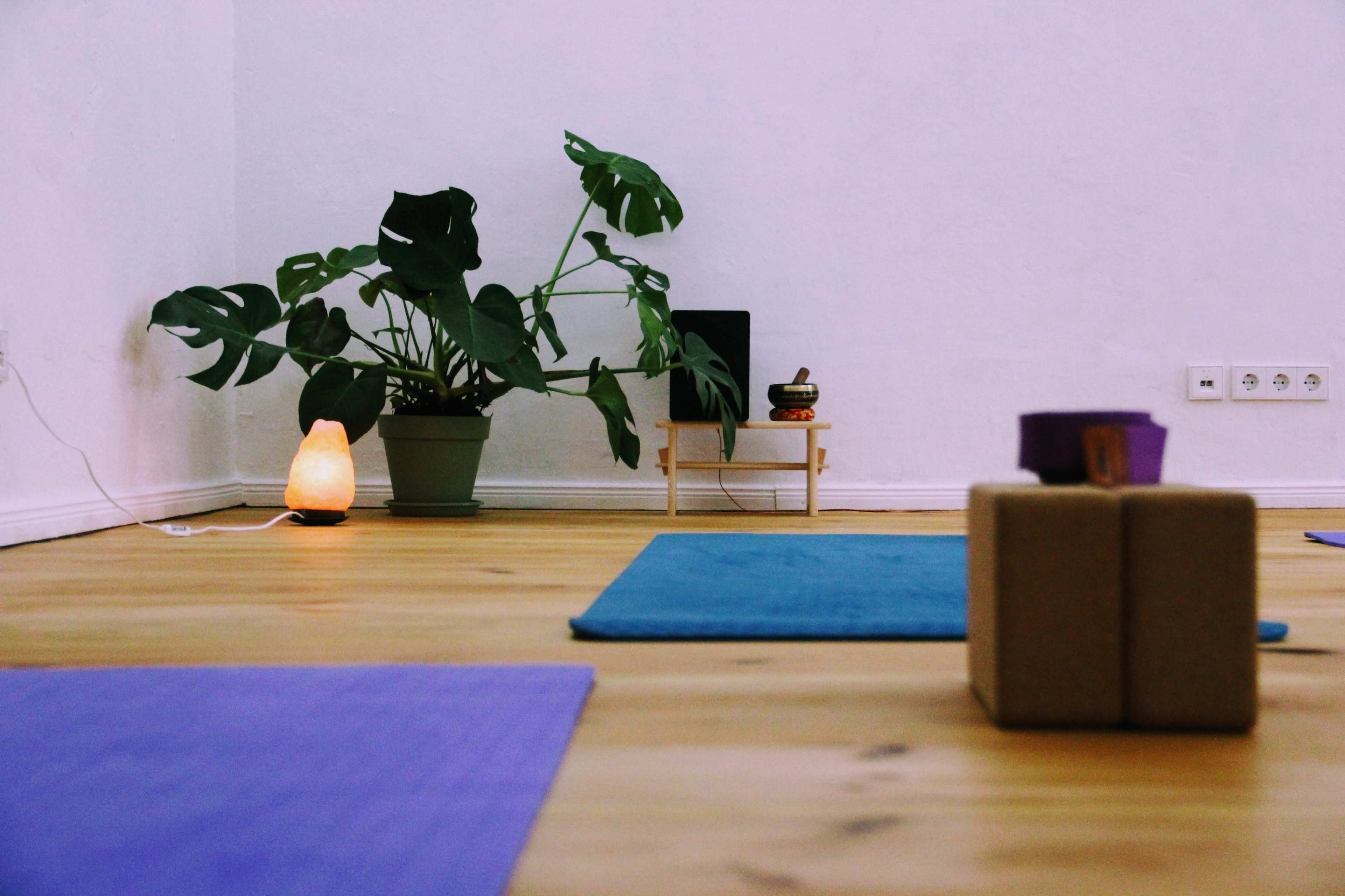 The image shows a minimalist yoga studio with exercise mats, a potted plant, and a softly lit lamp in the corner.