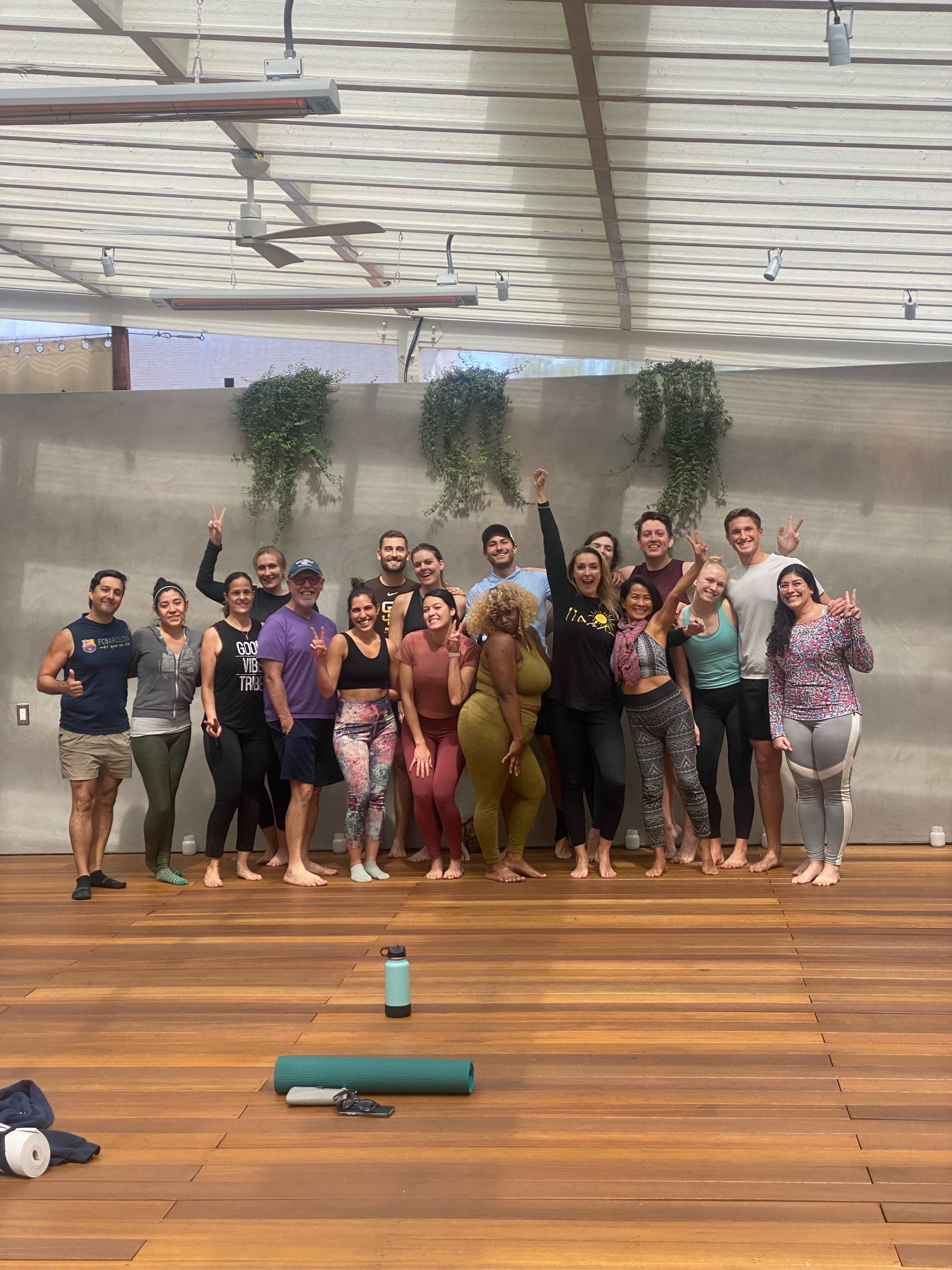 A diverse group of individuals poses together on a wooden yoga studio floor, smiling and making various peace signs.