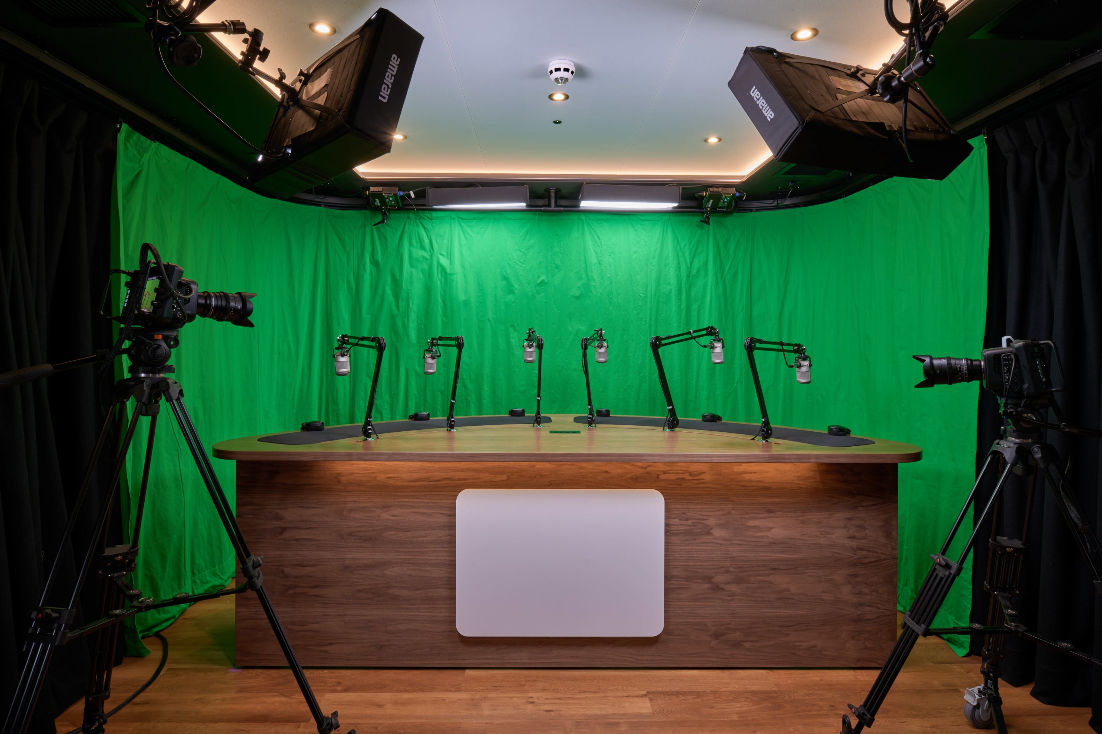 The image shows a studio setup with a wooden table, multiple microphone stands, and cameras arranged around a green backdrop.