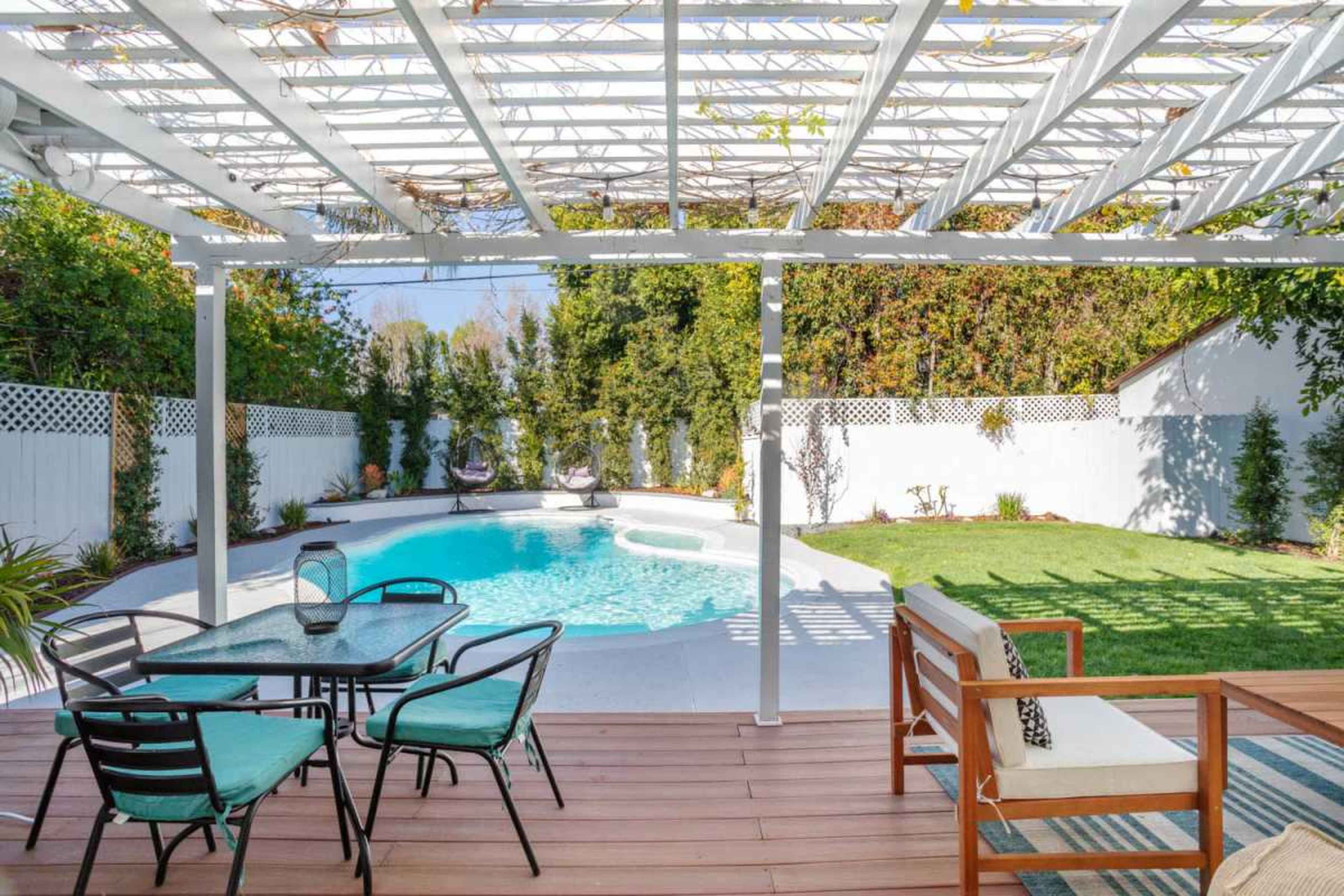The image shows a backyard with a swimming pool, surrounded by greenery and a shaded seating area with a dining set.