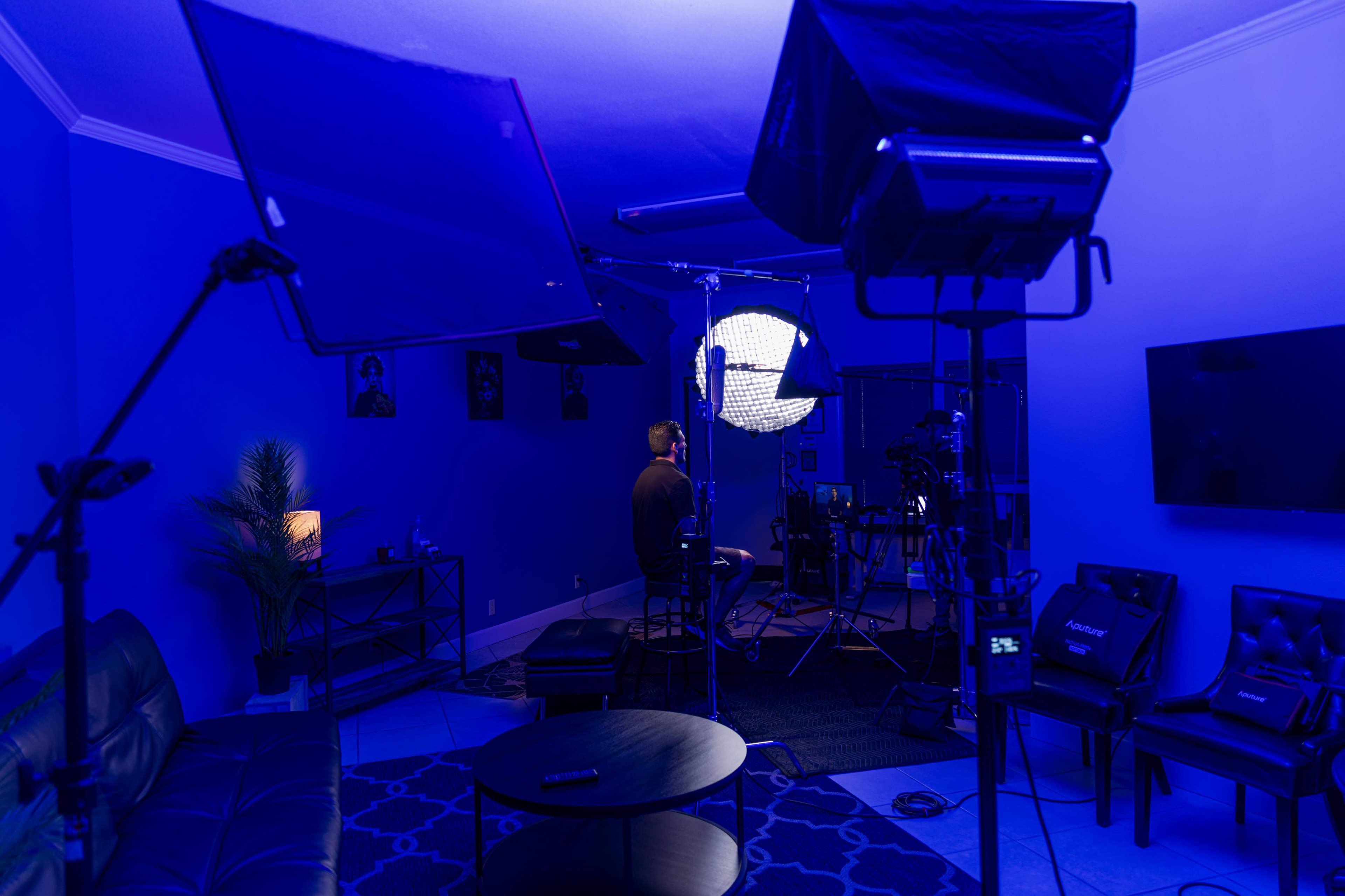 A person sits on a stool in a dimly lit studio space surrounded by photography lights and equipment.