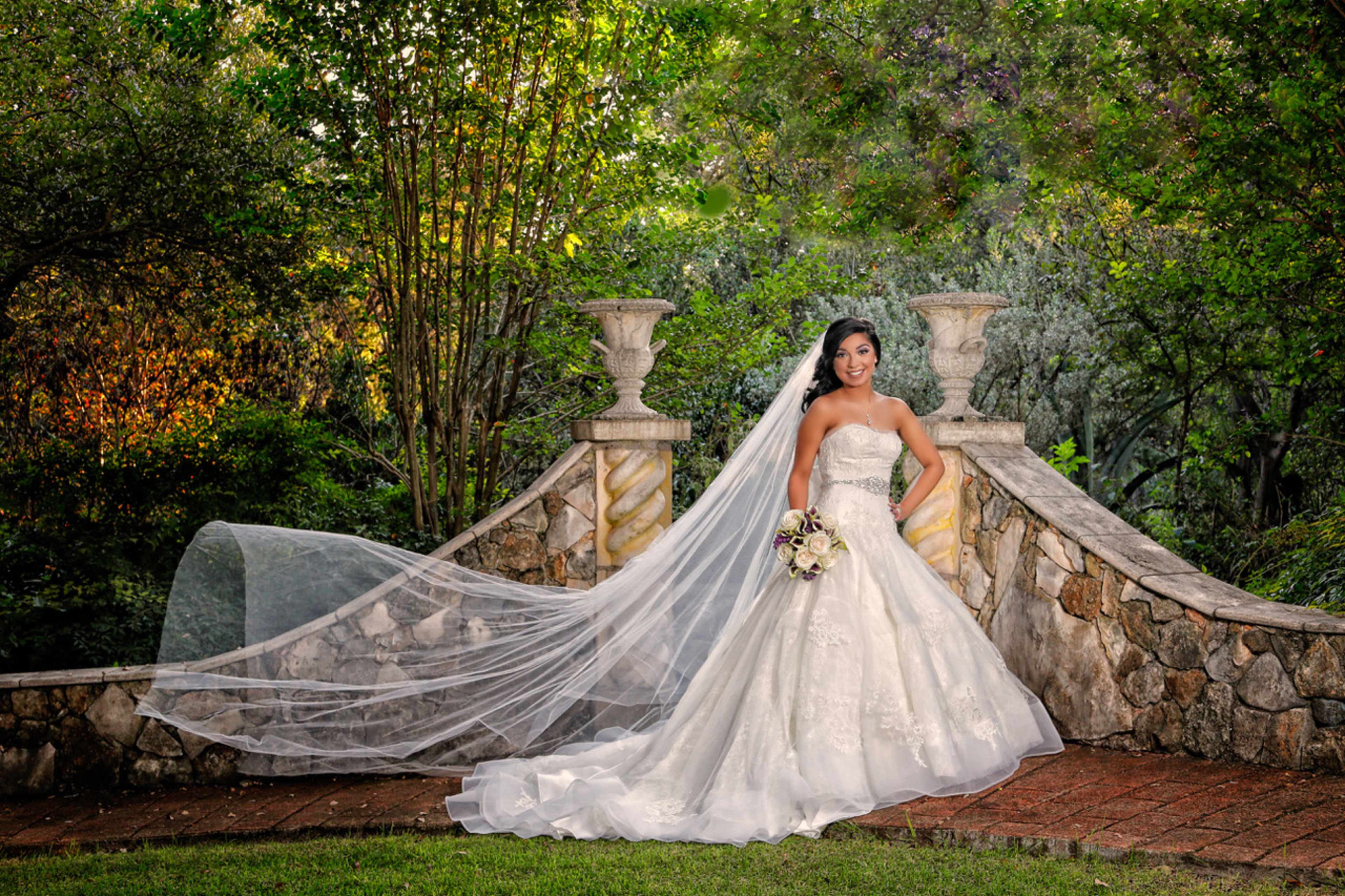 A bride in a white gown with a long veil stands on a stone pathway surrounded by greenery.