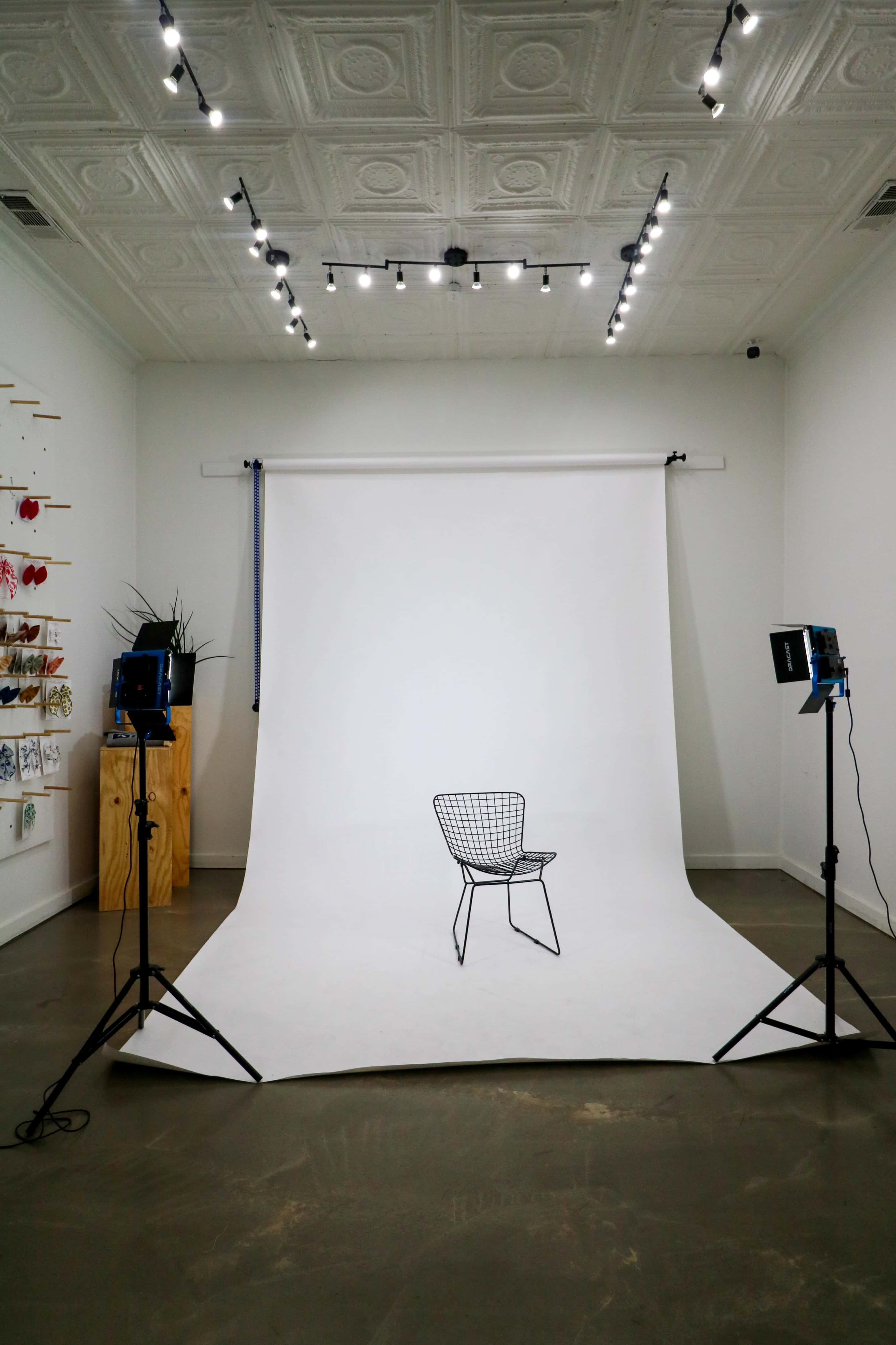 A minimalist photography studio features a wire chair positioned in front of a white backdrop, illuminated by two softbox lights.