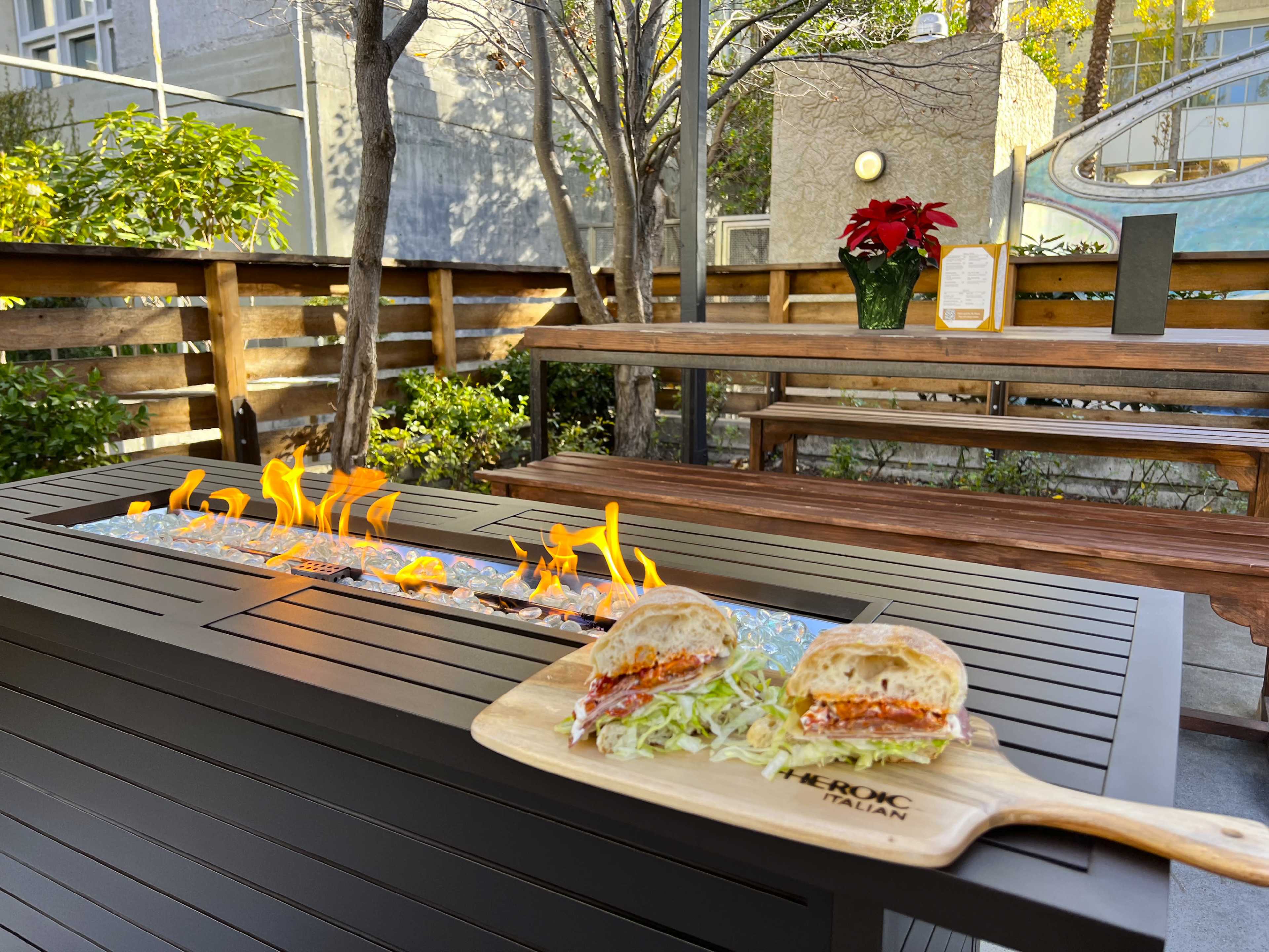 A sandwich is placed on a wooden cutting board beside a fire pit surrounded by benches in a landscaped outdoor setting.
