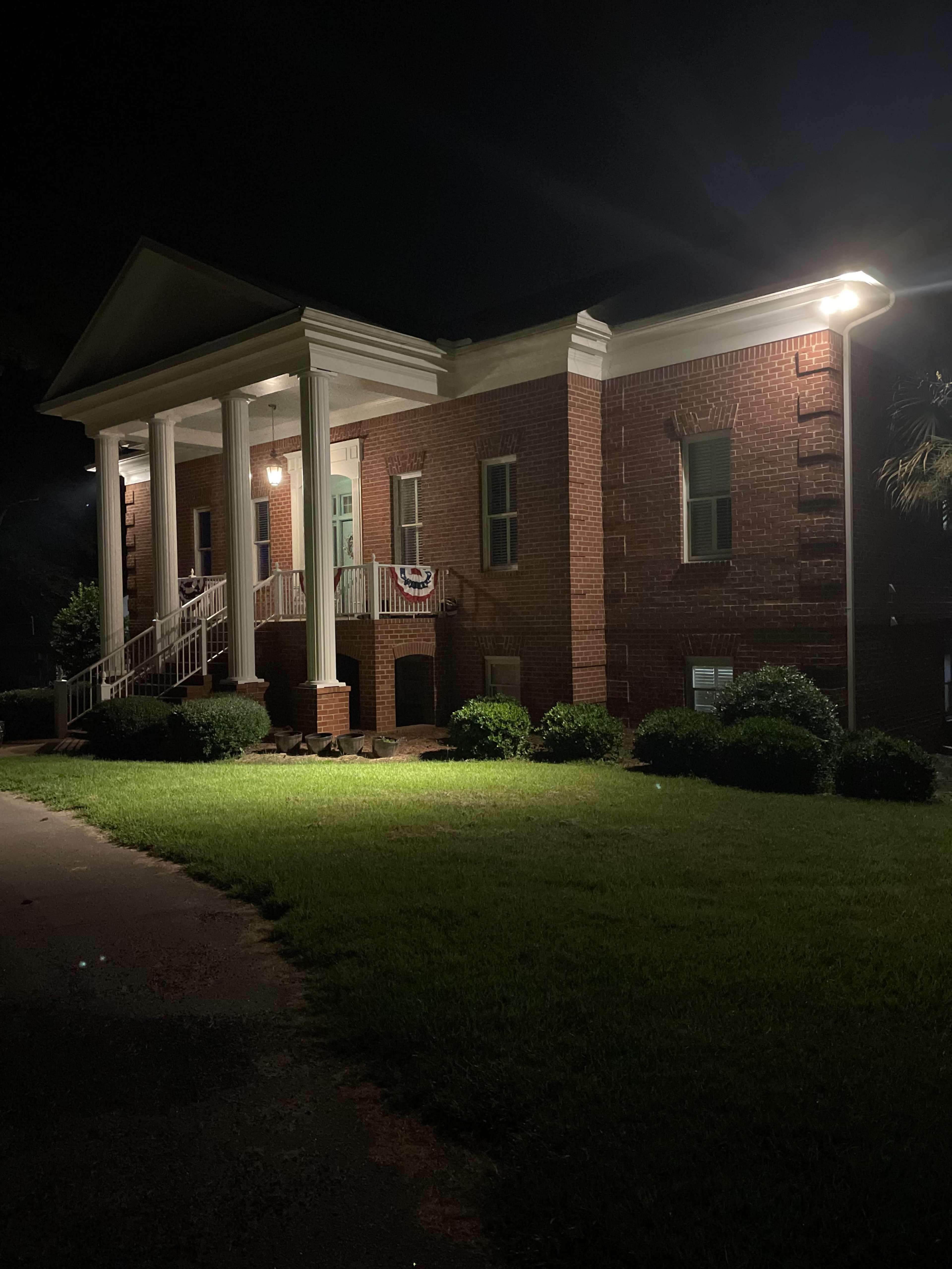 A two-story red brick building with white columns is illuminated by outdoor lighting at night.