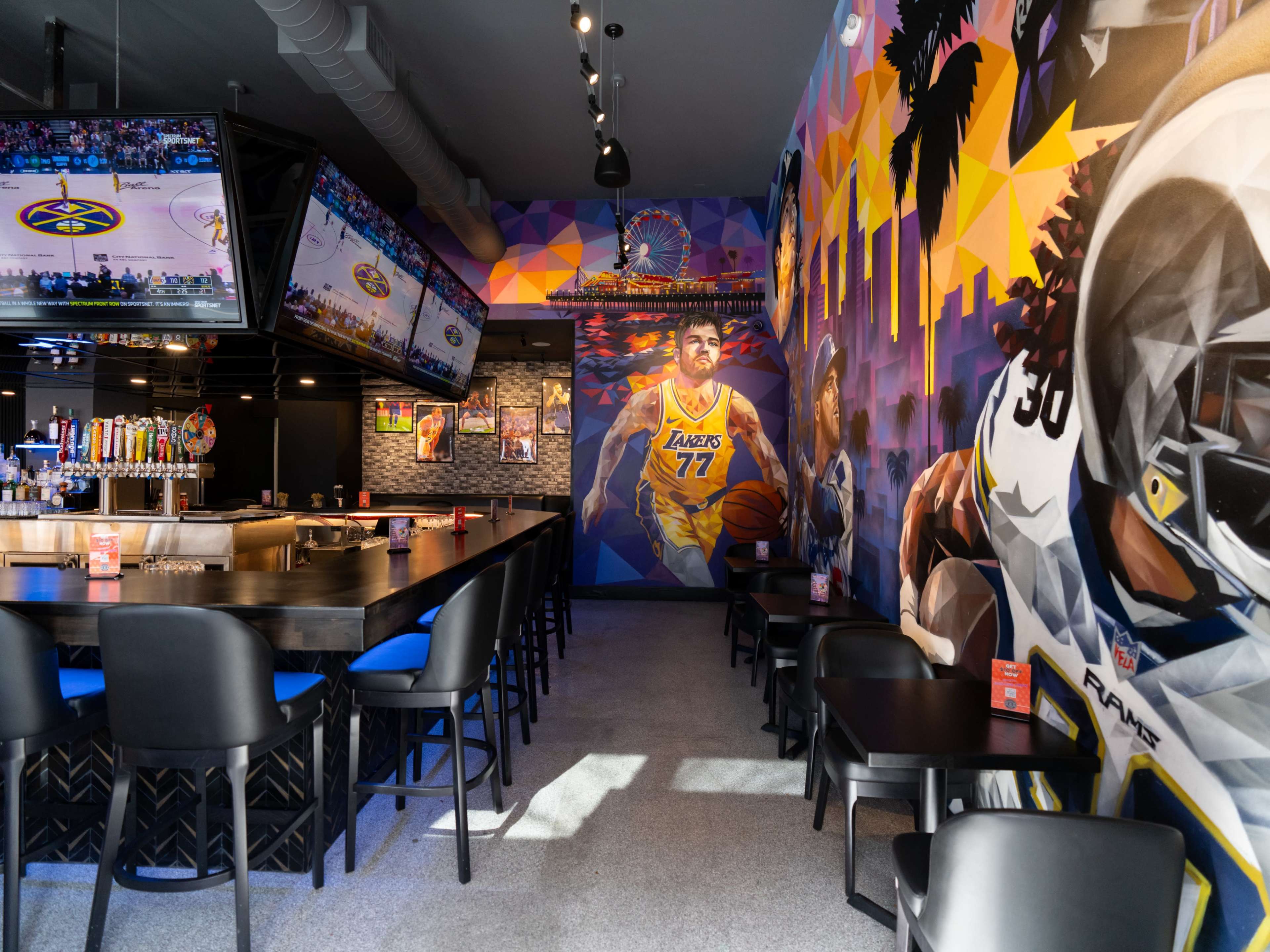 The image shows a sports bar interior featuring a mural of a basketball player, televisions displaying a game, and black tables and chairs.