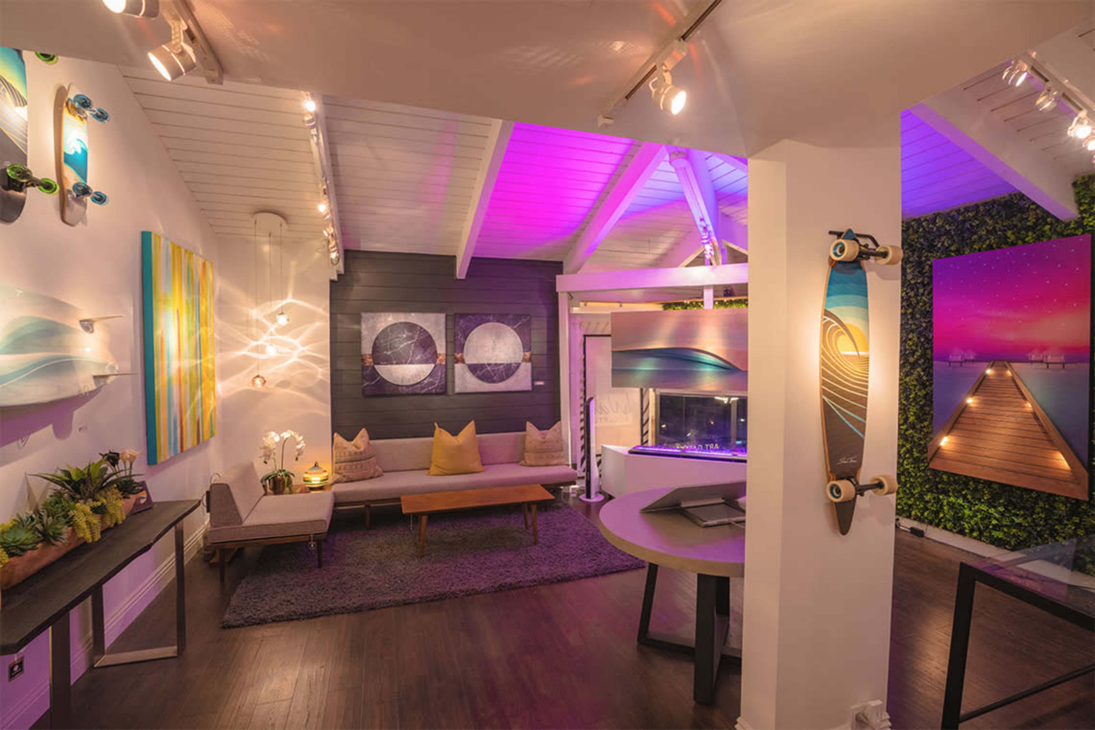 The image shows a modern indoor space with artwork on the walls, a cozy seating area, and vibrant lighting.