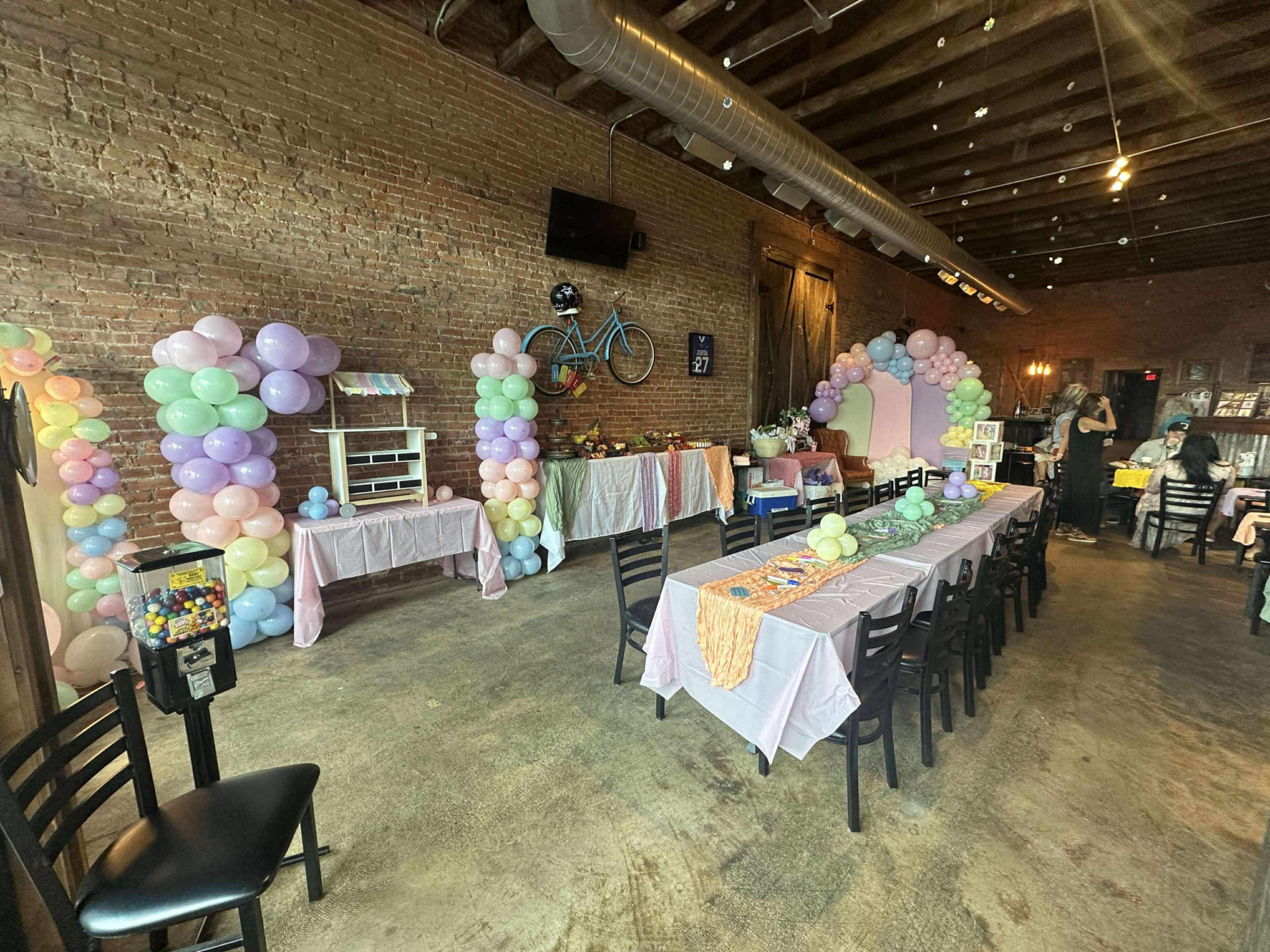 The image shows a party venue with tables dressed in light pink coverings and decorated with pastel balloons arranged in arches along the walls.
