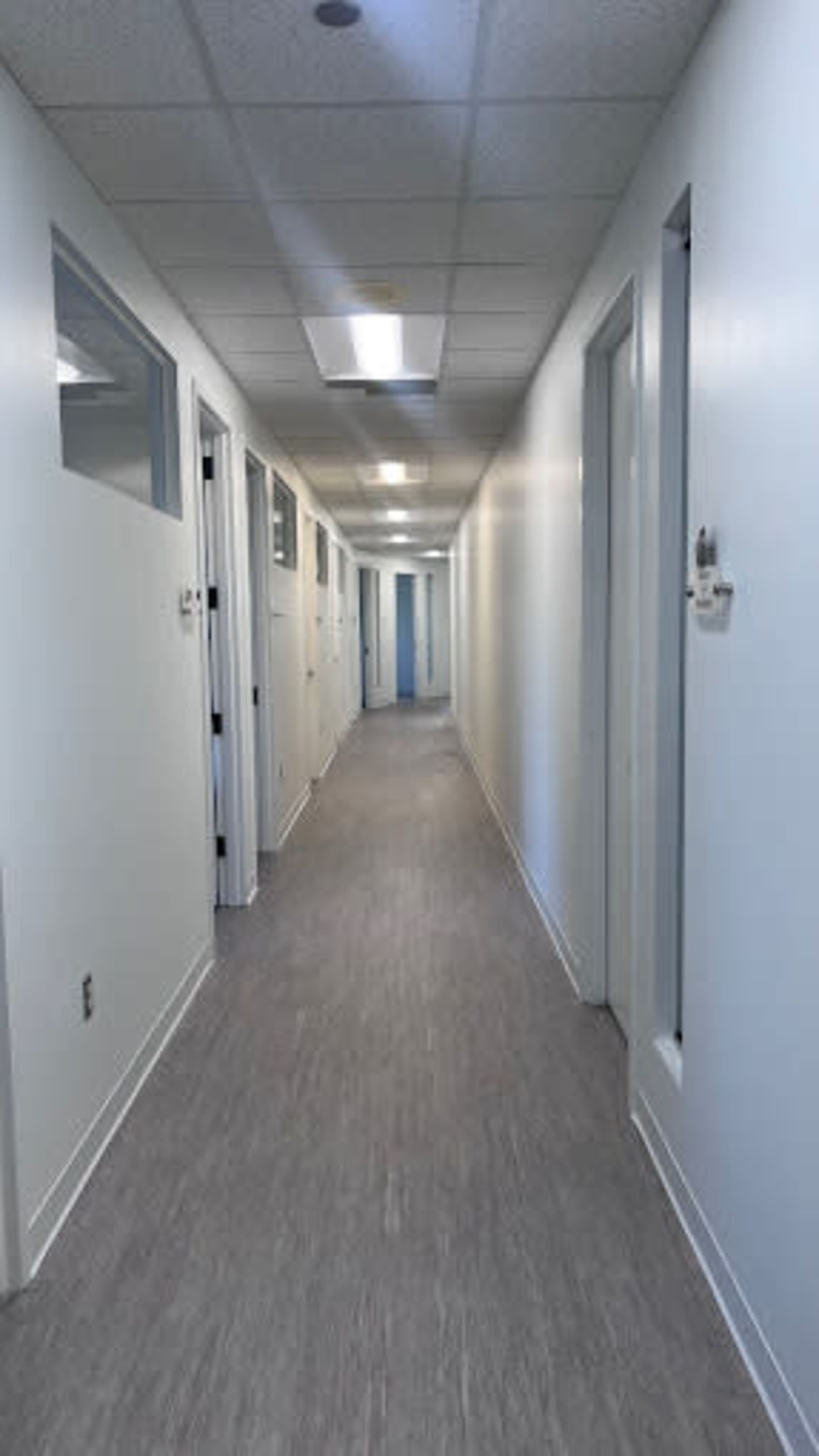 The image shows a long, narrow hallway lined with white doors and illuminated by overhead lights.