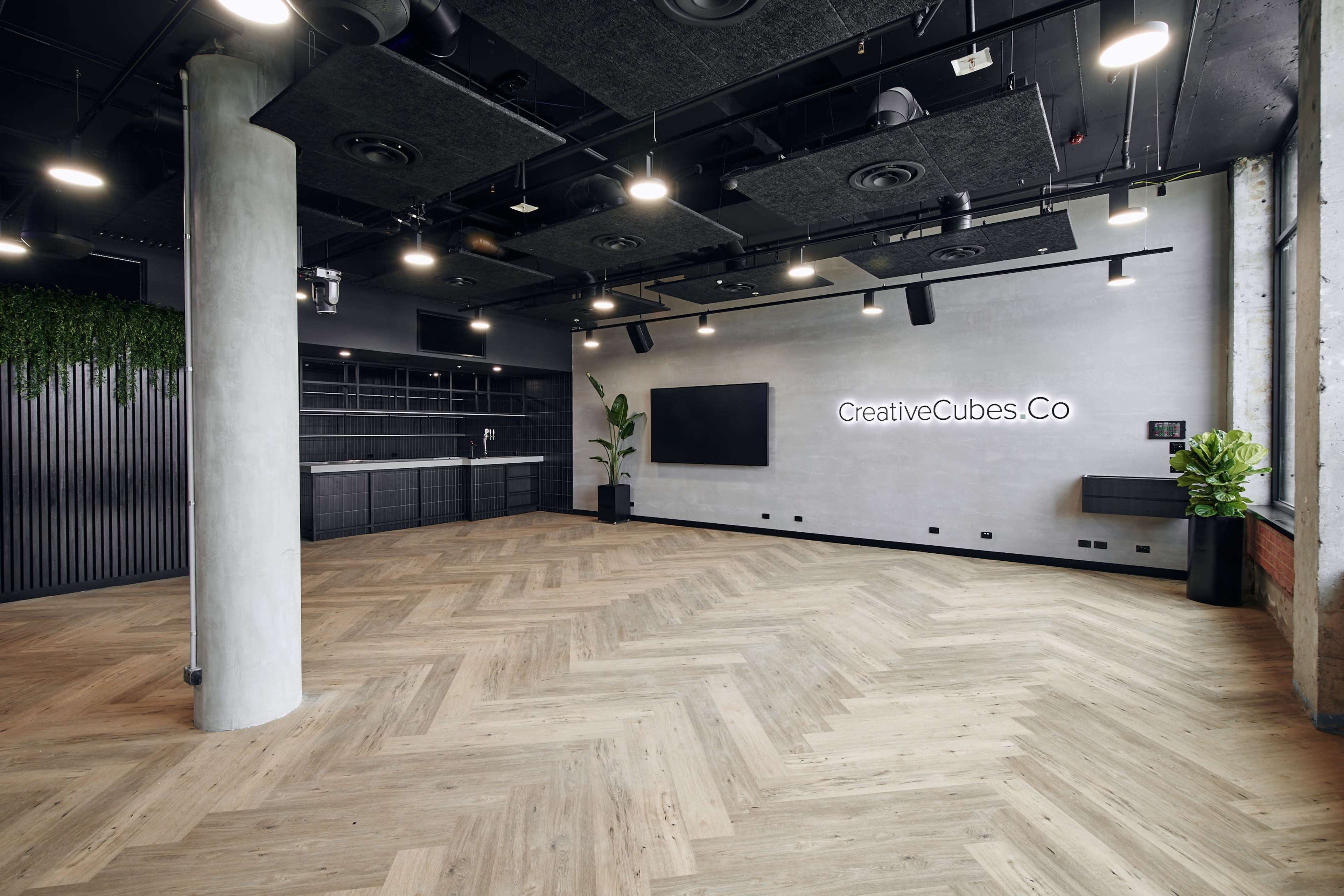 The image shows a modern, empty office space with wooden herringbone flooring, concrete columns, and a large wall-mounted screen surrounded by sleek cabinetry and plants.