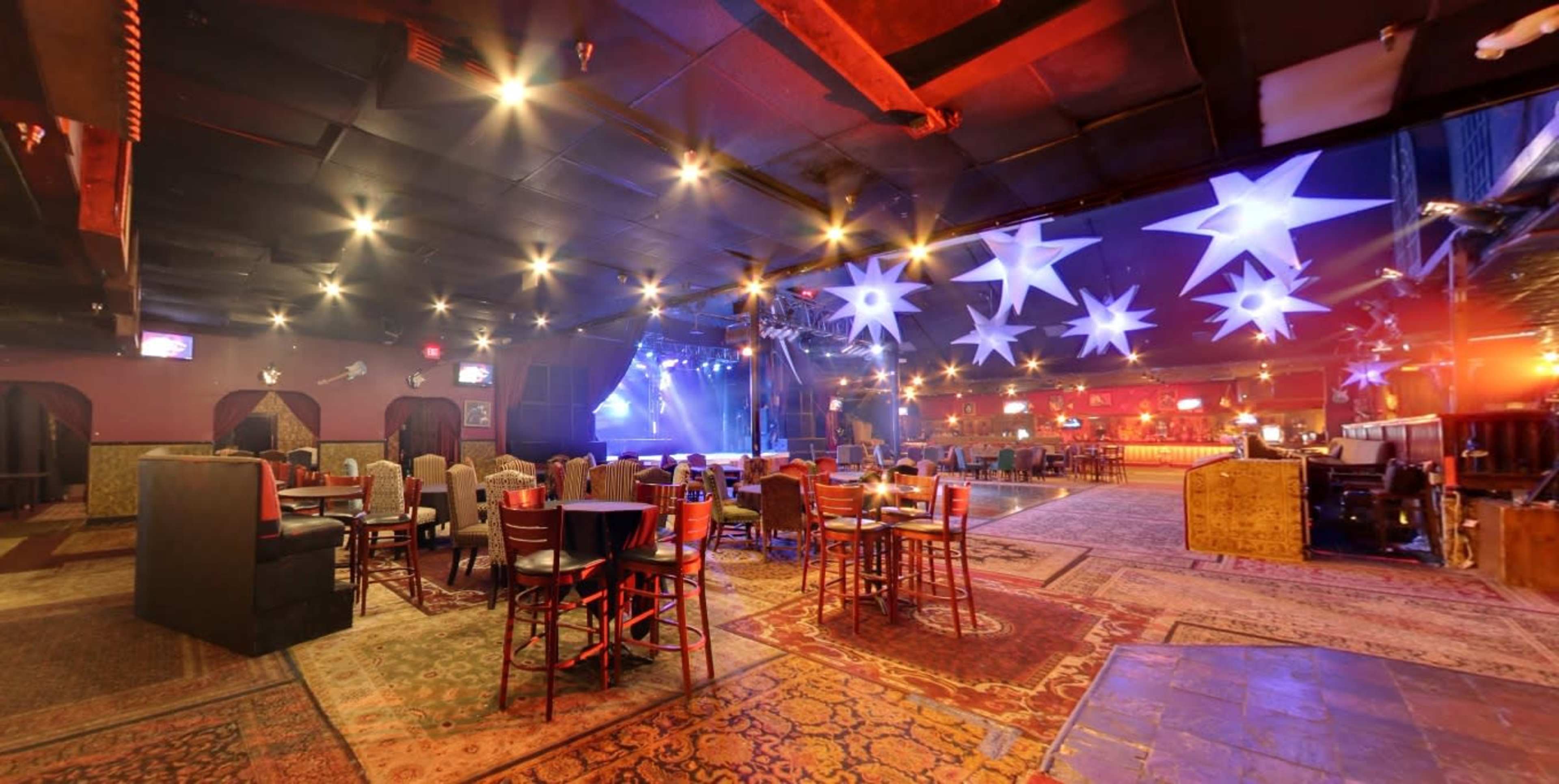 A spacious event venue features rows of wooden chairs and tables, illuminated by colorful lights and adorned with hanging star-shaped decorations.