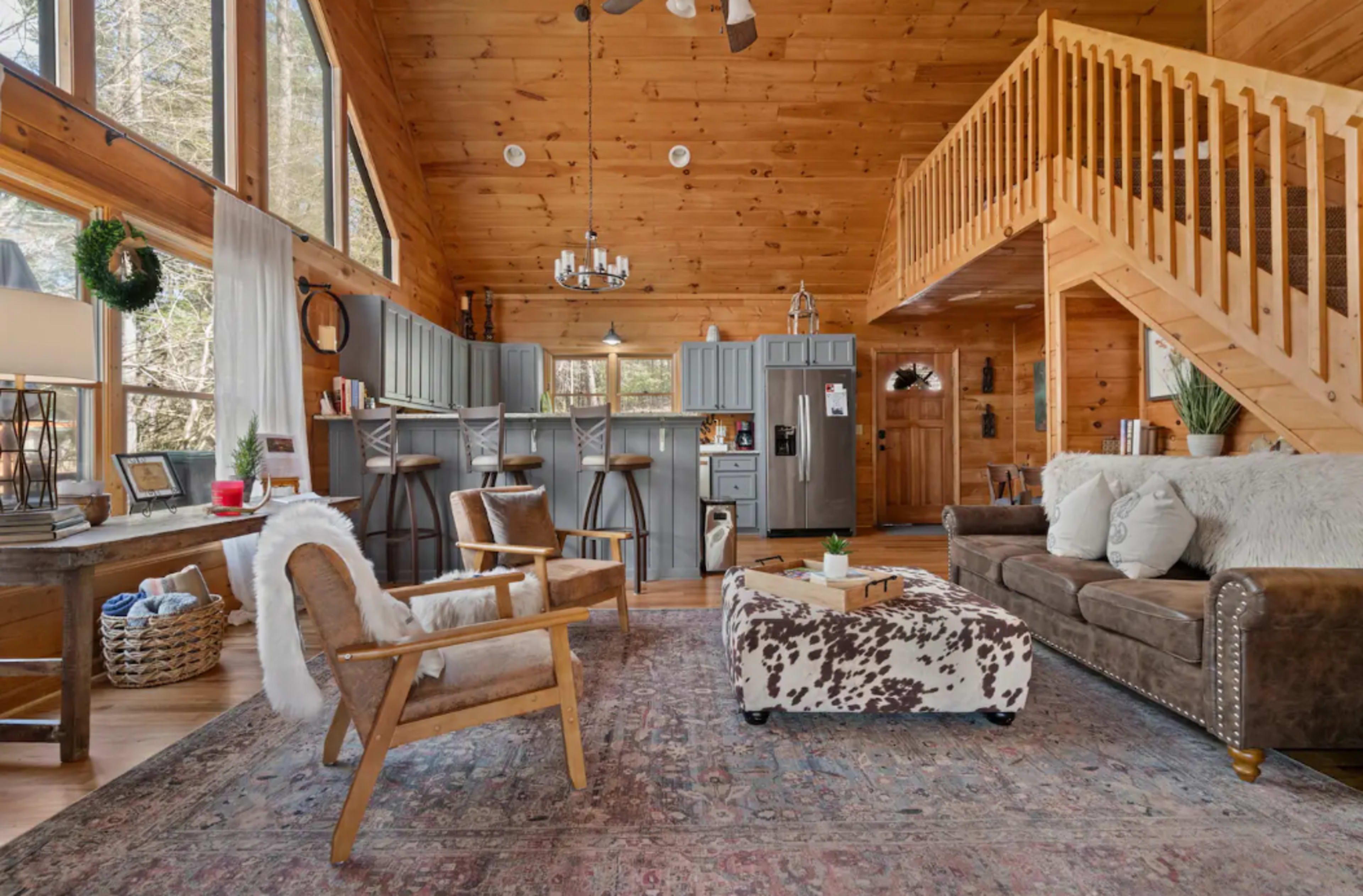 A spacious, wooden cabin interior featuring a living area with a brown sofa, armchairs, and a patterned ottoman, alongside a staircase and a kitchen area with bar seating.