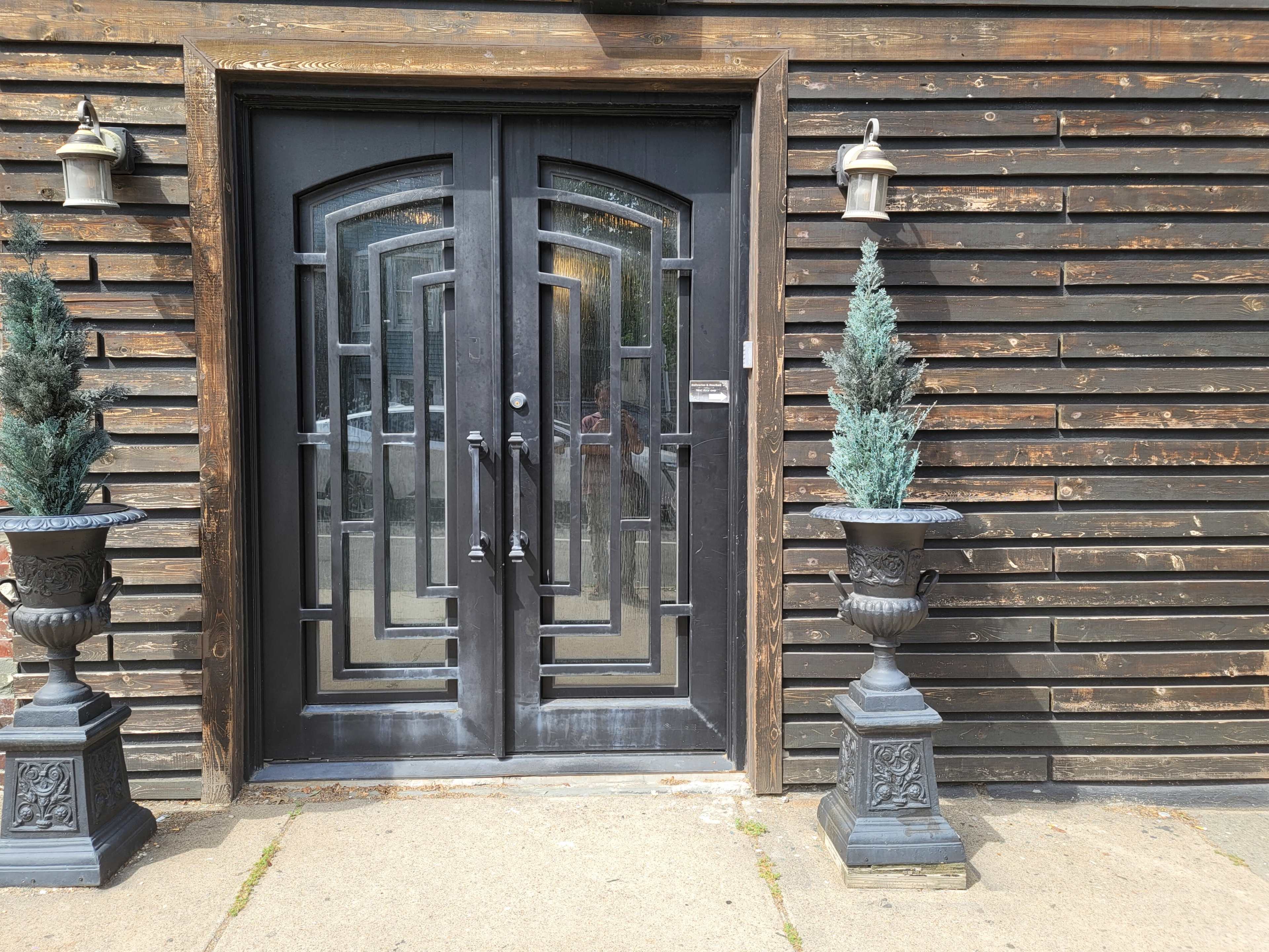The image shows a pair of black double doors with decorative glass panels, flanked by two large planters containing coniferous plants, set against a wooden wall.