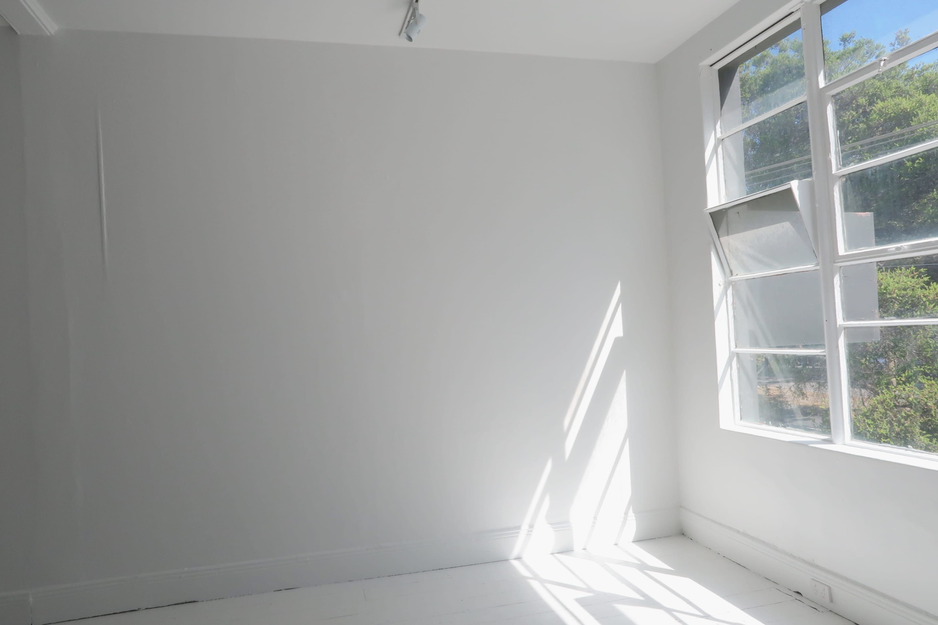 A bright, empty room features white walls and a large window allowing sunlight to stream in.
