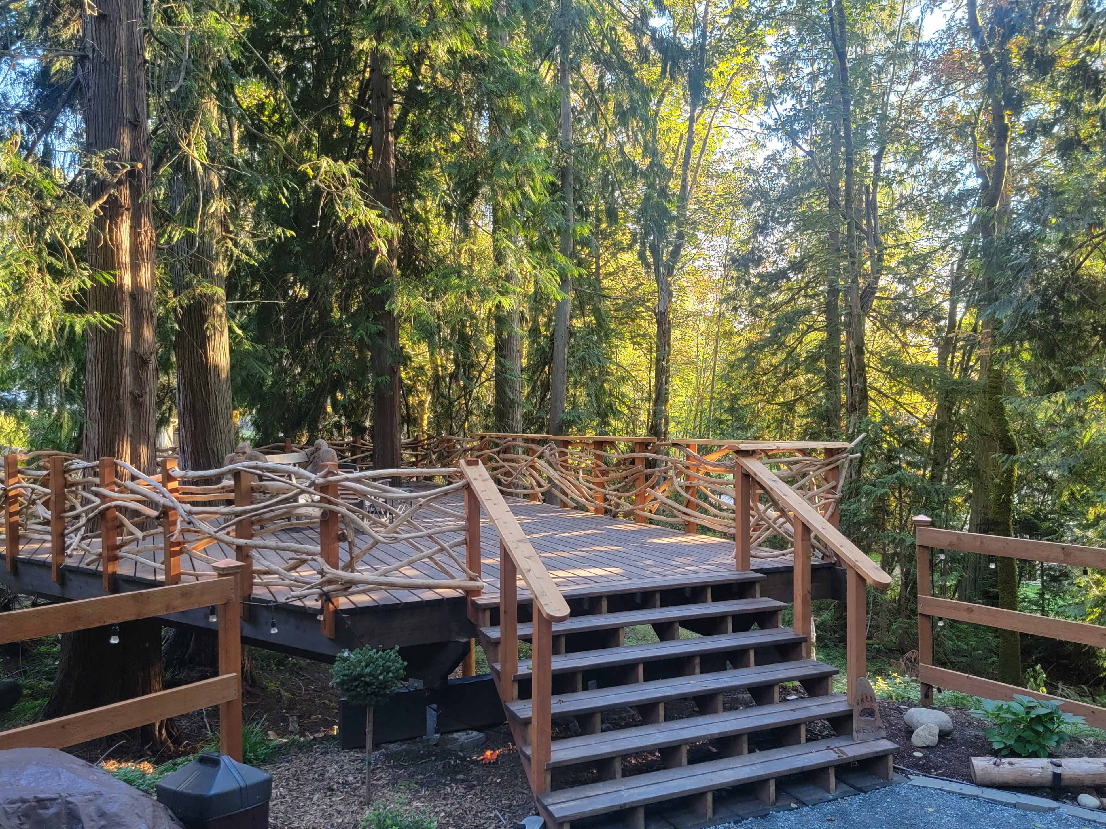 The image shows a wooden deck surrounded by tall trees in a forested area, featuring a railing made from branches and a set of stairs leading up to it.