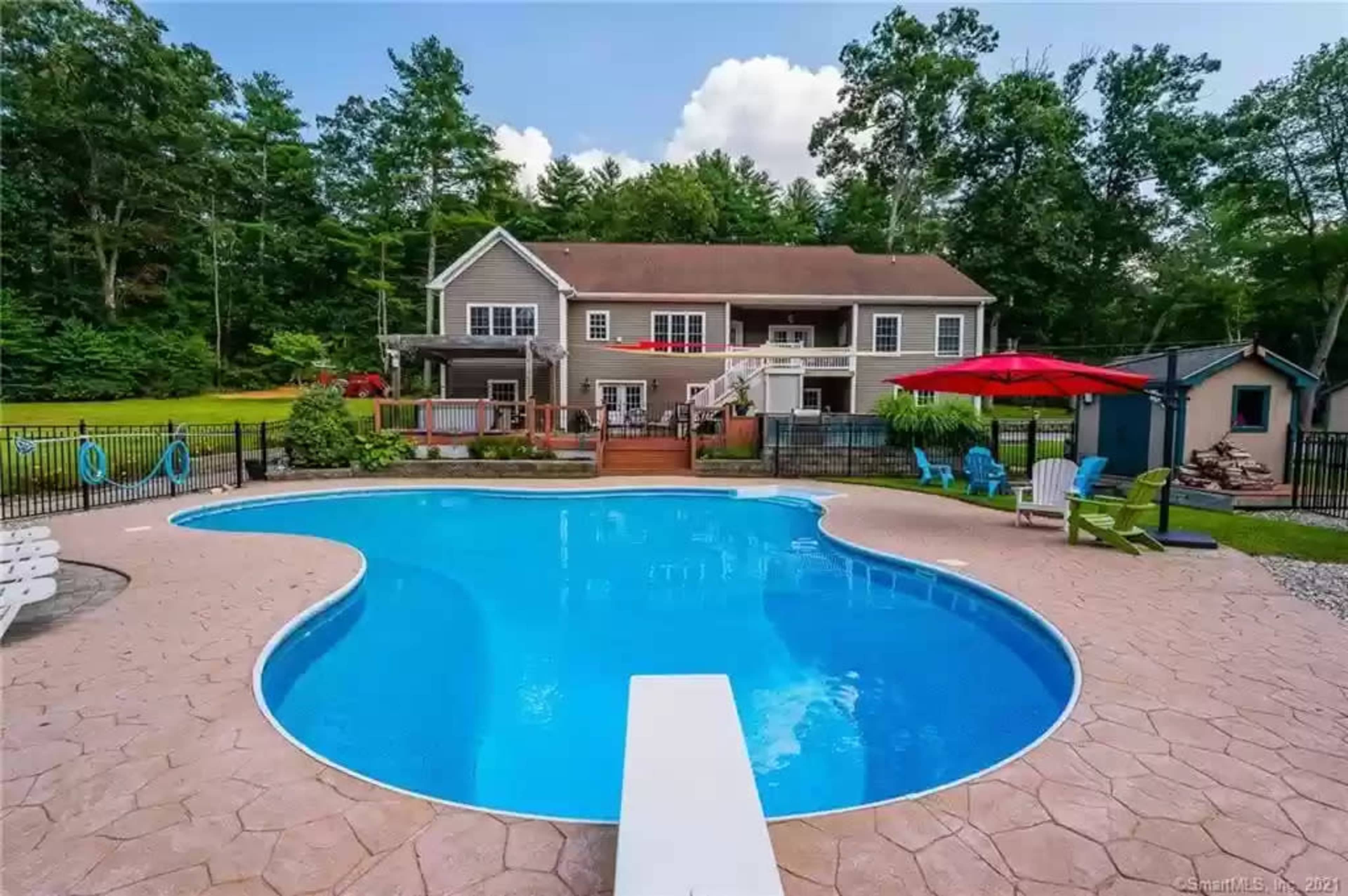 A backyard swimming pool with a diving board, surrounded by a patio and green trees, with a house in the background.