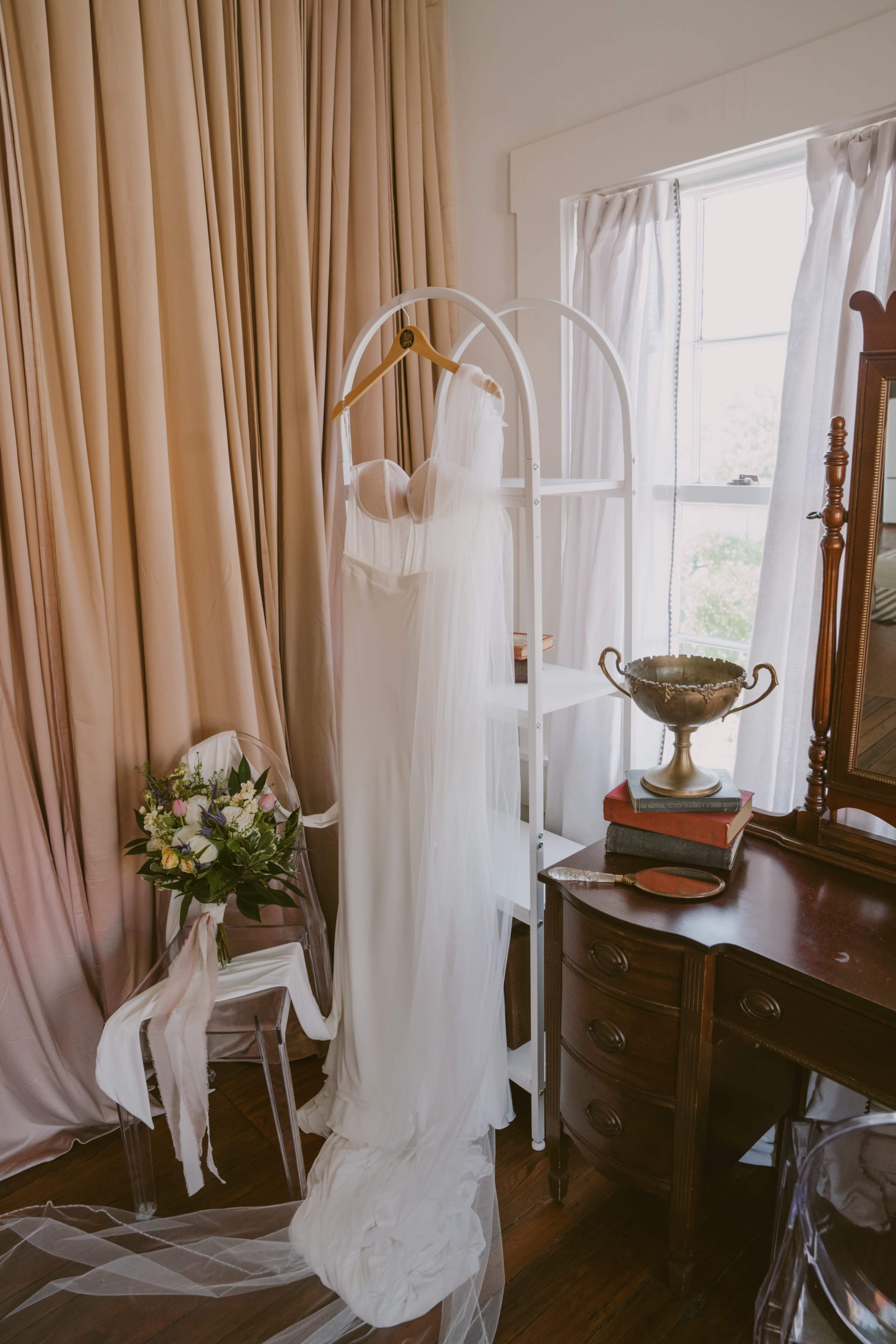 A white wedding dress is hanging on a display rack next to a bouquet of flowers and a vintage wooden dresser with a decorative trophy.