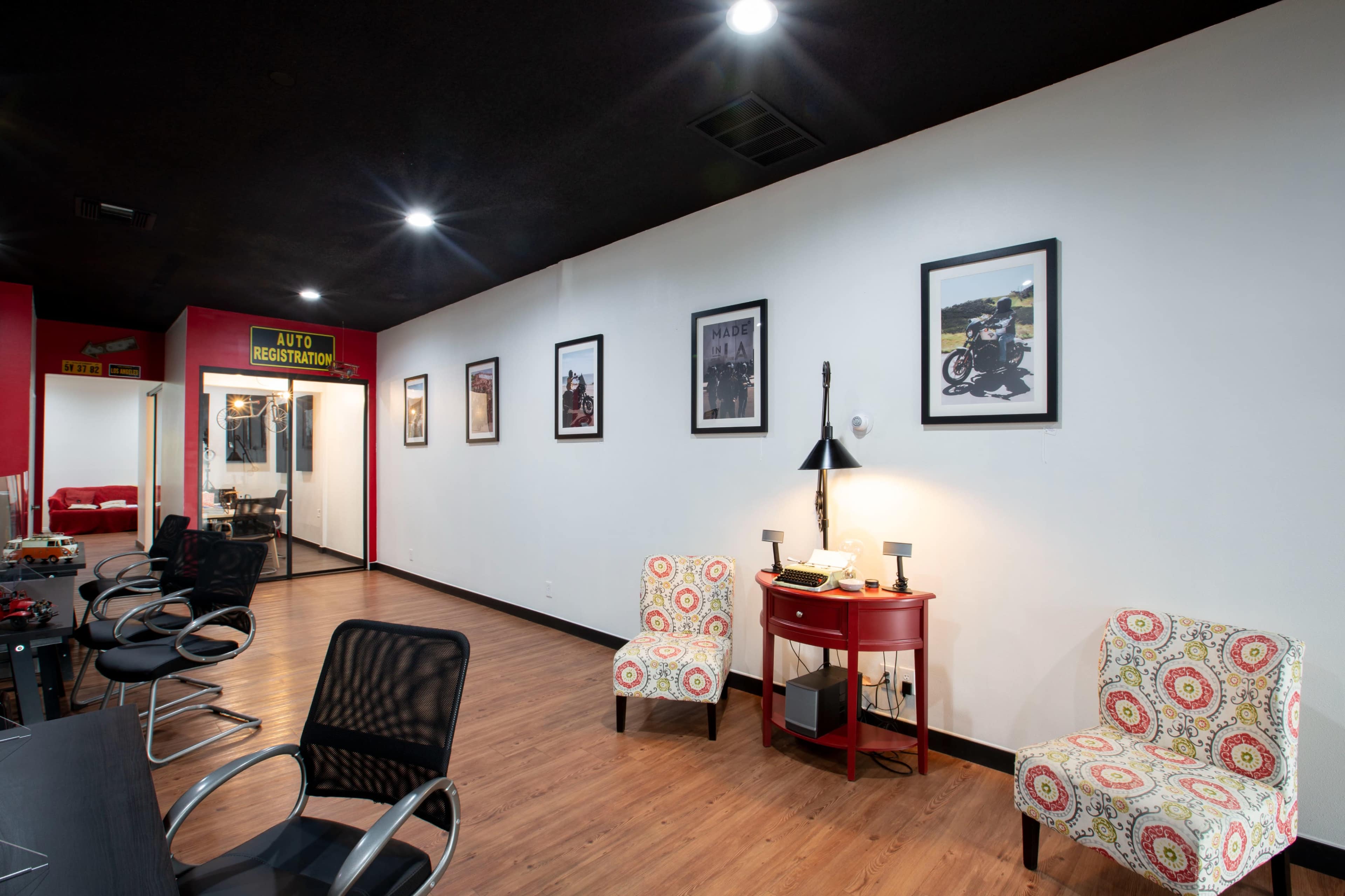 The image shows a modern waiting area with two patterned chairs, a red side table, and black office chairs lining the left side against white walls adorned with framed photographs.