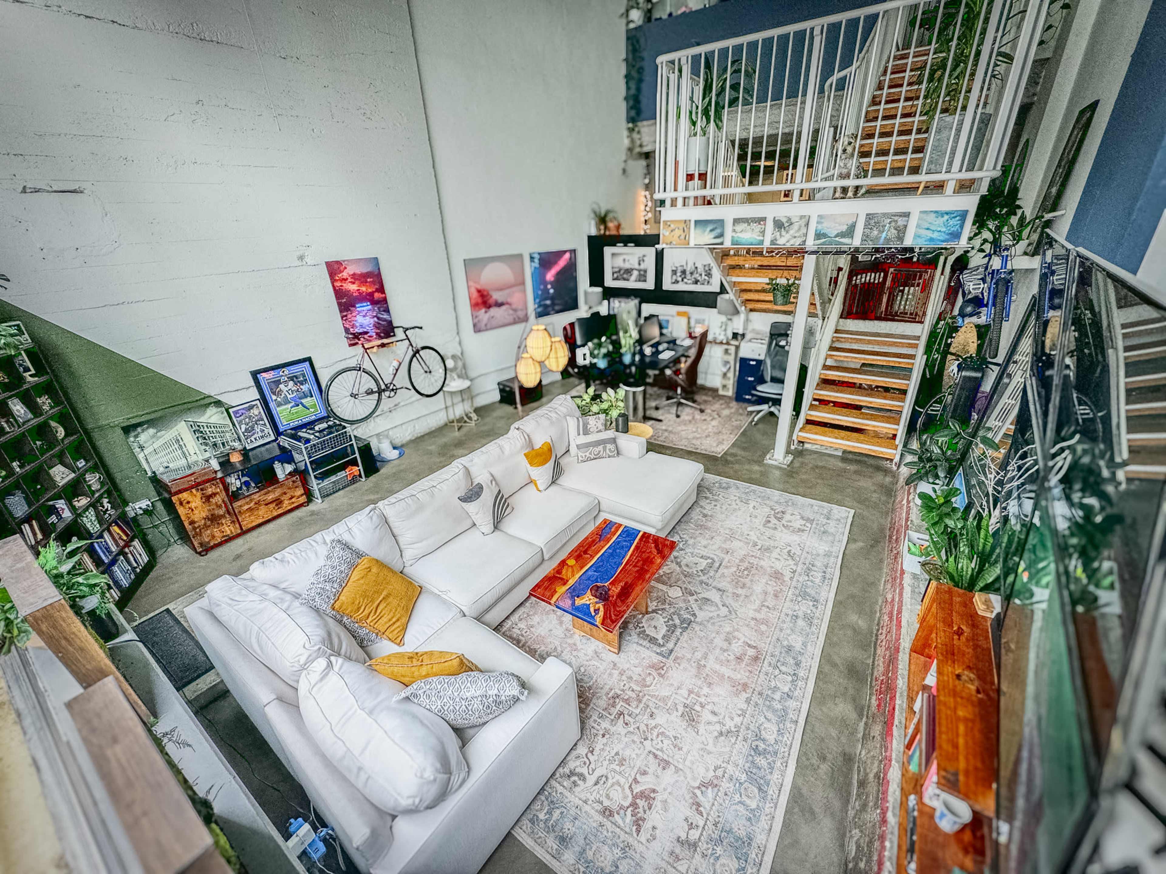 A spacious, modern loft-style living area featuring a large white sectional sofa, a wooden coffee table, and a staircase leading to an upper level decorated with plants and artwork.