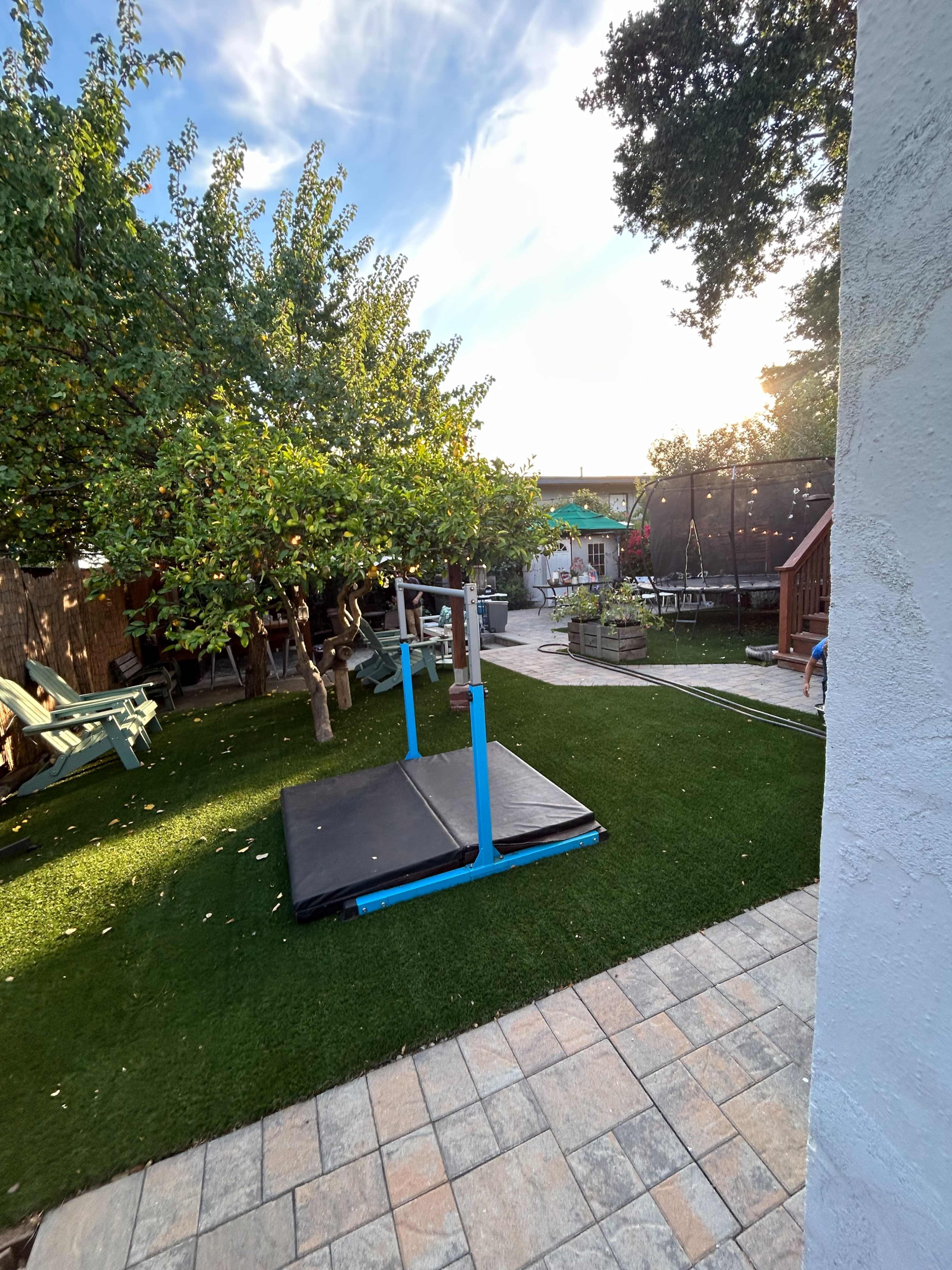 A backyard features a grassy area with a gymnastics mat, trees, and a patio, alongside various outdoor seating and a playground.