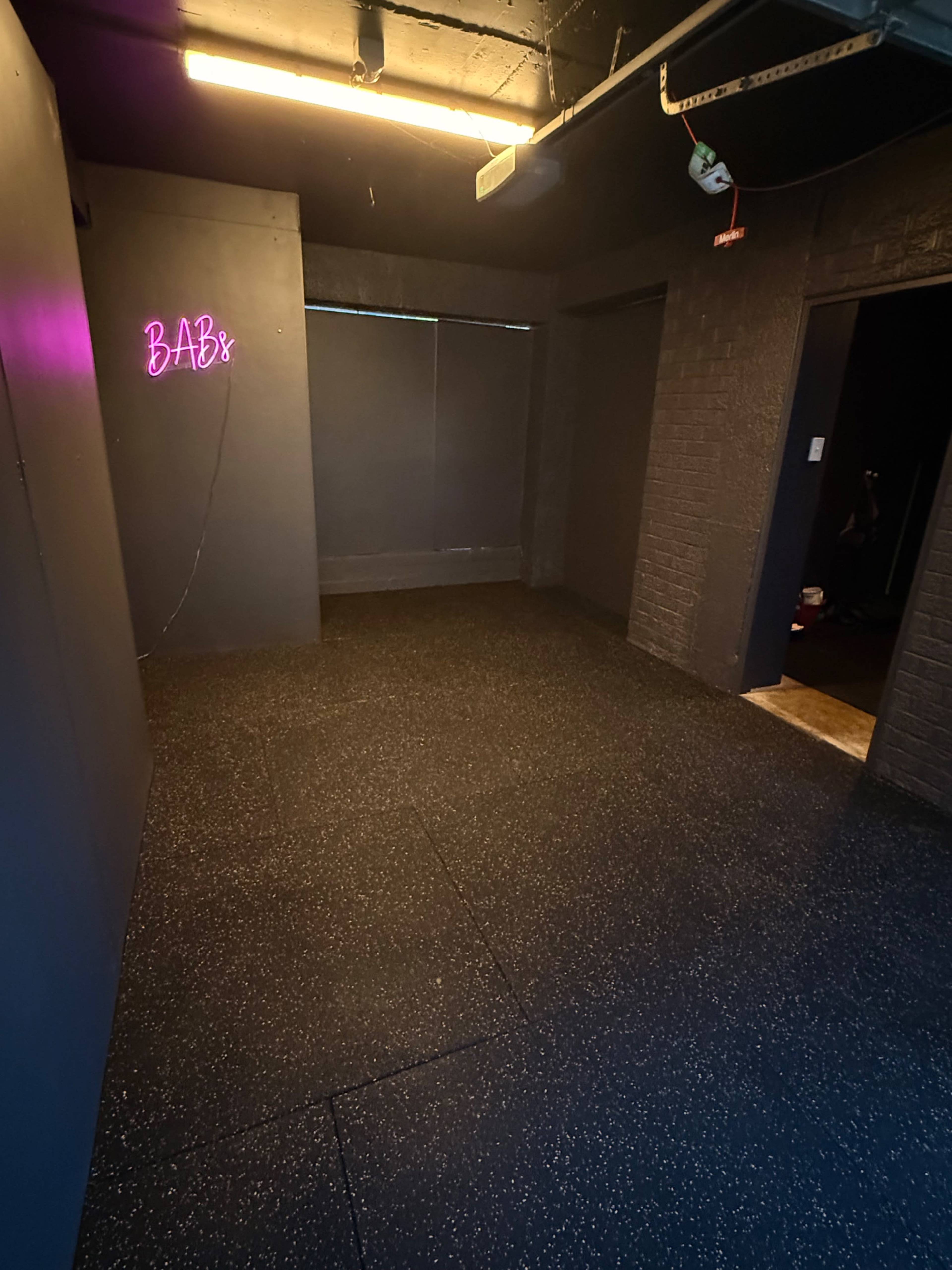 A dimly lit room features black walls, a rubber floor, and a neon sign that says "BABE" on one side.