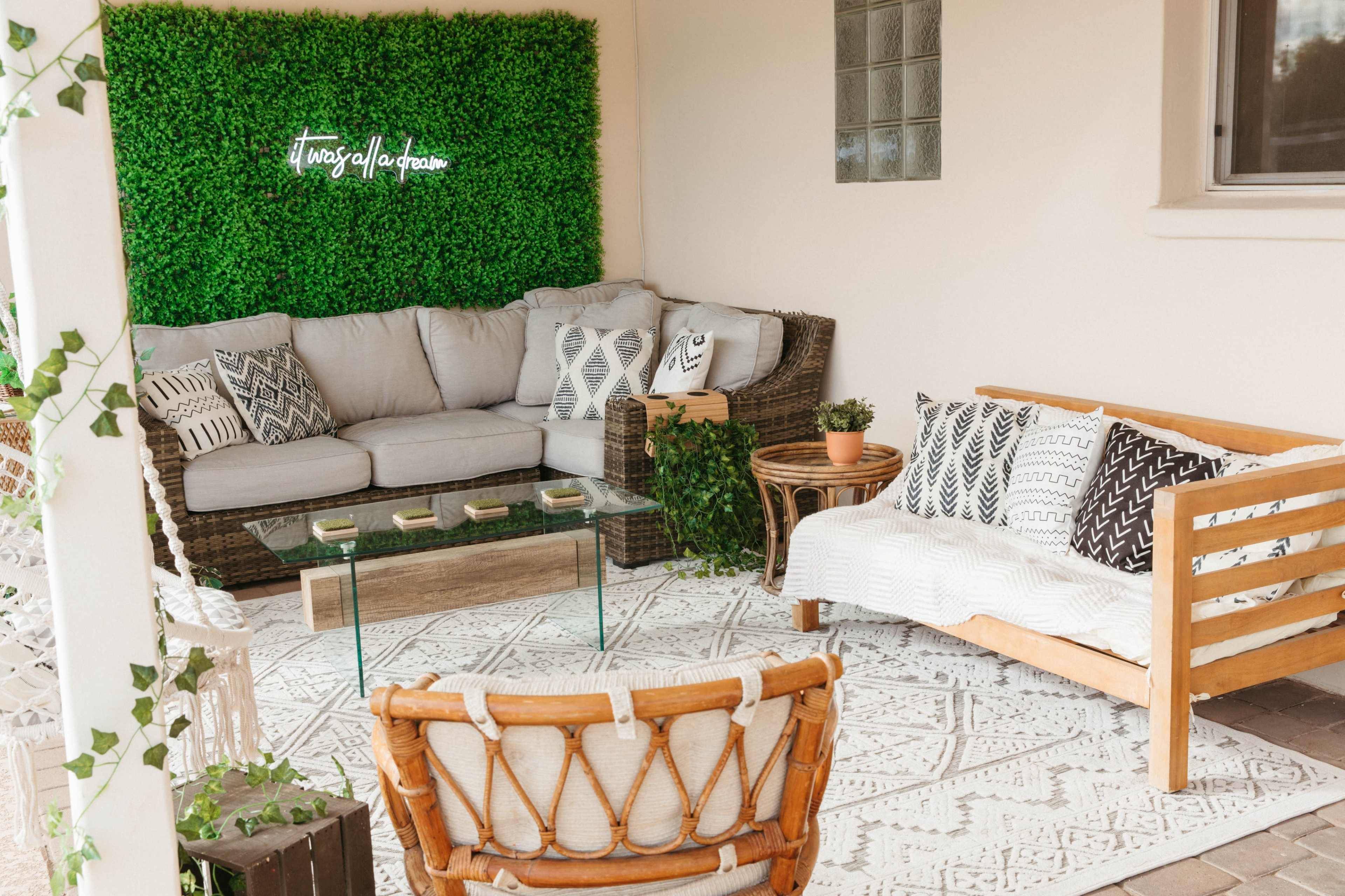A cozy outdoor seating area features a sofa and chairs arranged around a glass coffee table, with a backdrop of greenery and decorative pillows.