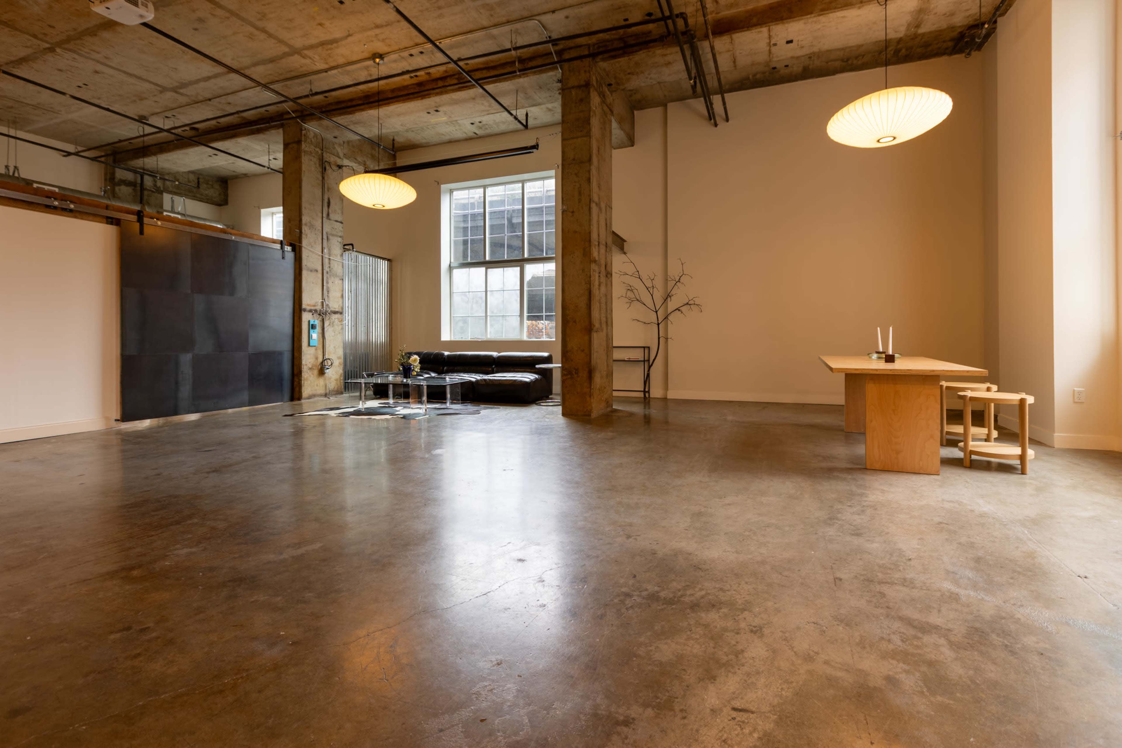 The image shows a spacious, open-concept interior with concrete floors, large windows, a black leather sofa, a wooden dining table, and minimalist decor.