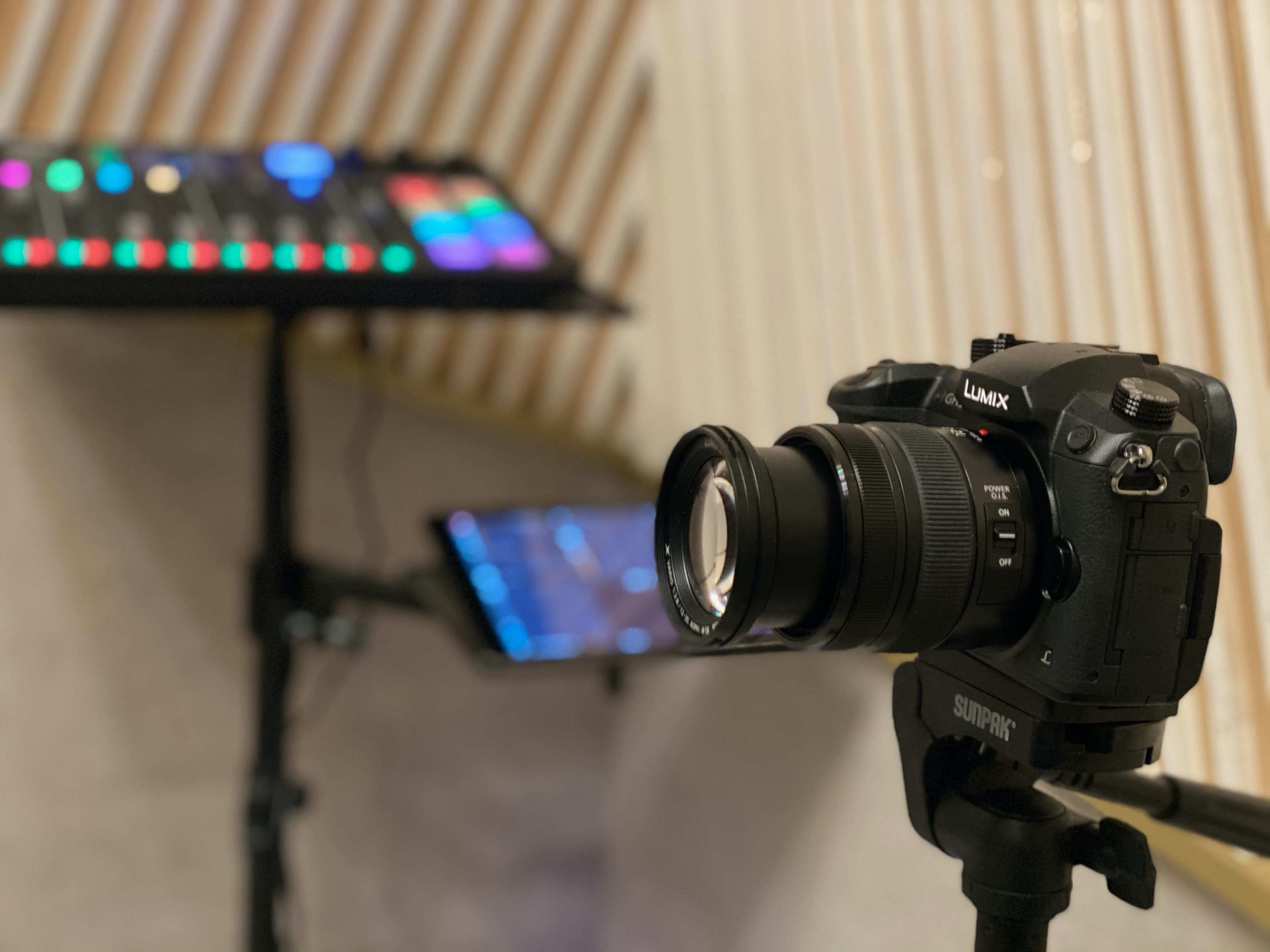 A camera is positioned on a tripod in front of a digital mixing board with colorful control lights.