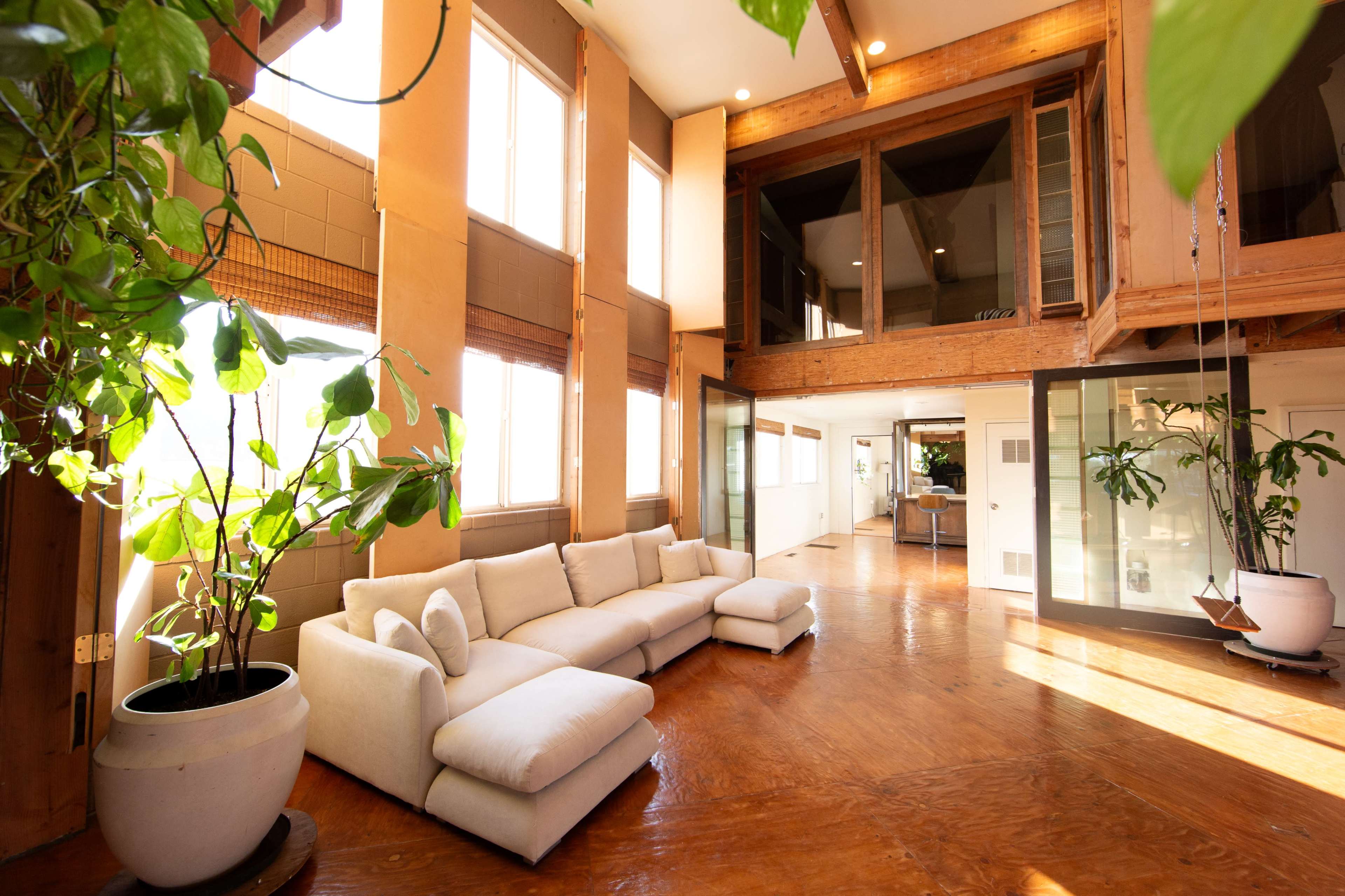 The image shows a spacious living area with large windows, a sectional sofa, and plants, featuring wooden beams and a contemporary design.