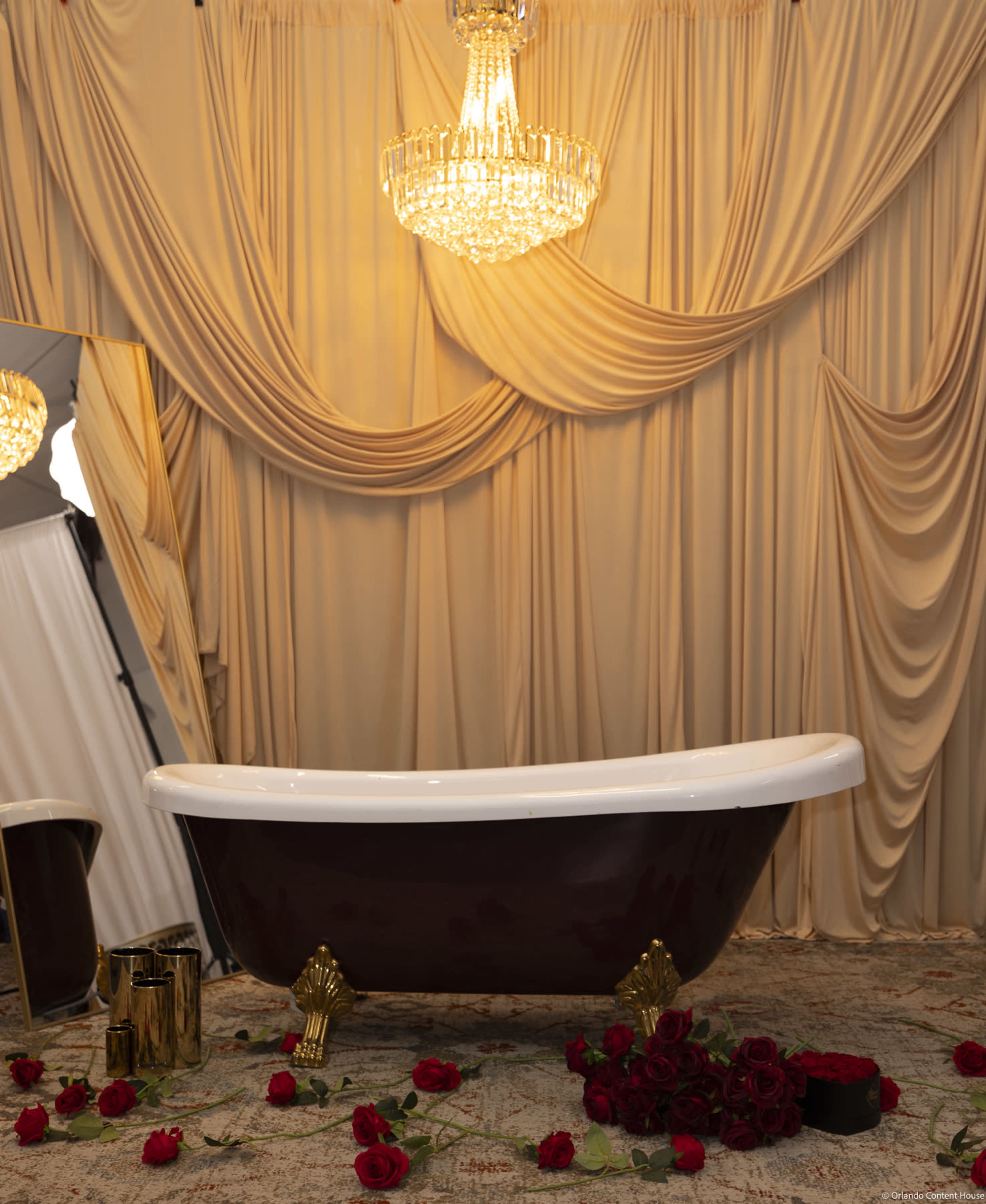 The image features a vintage-style bathtub with golden claw feet, positioned on a decorative carpet surrounded by scattered red roses, under a crystal chandelier against a backdrop of elegant draped fabric.