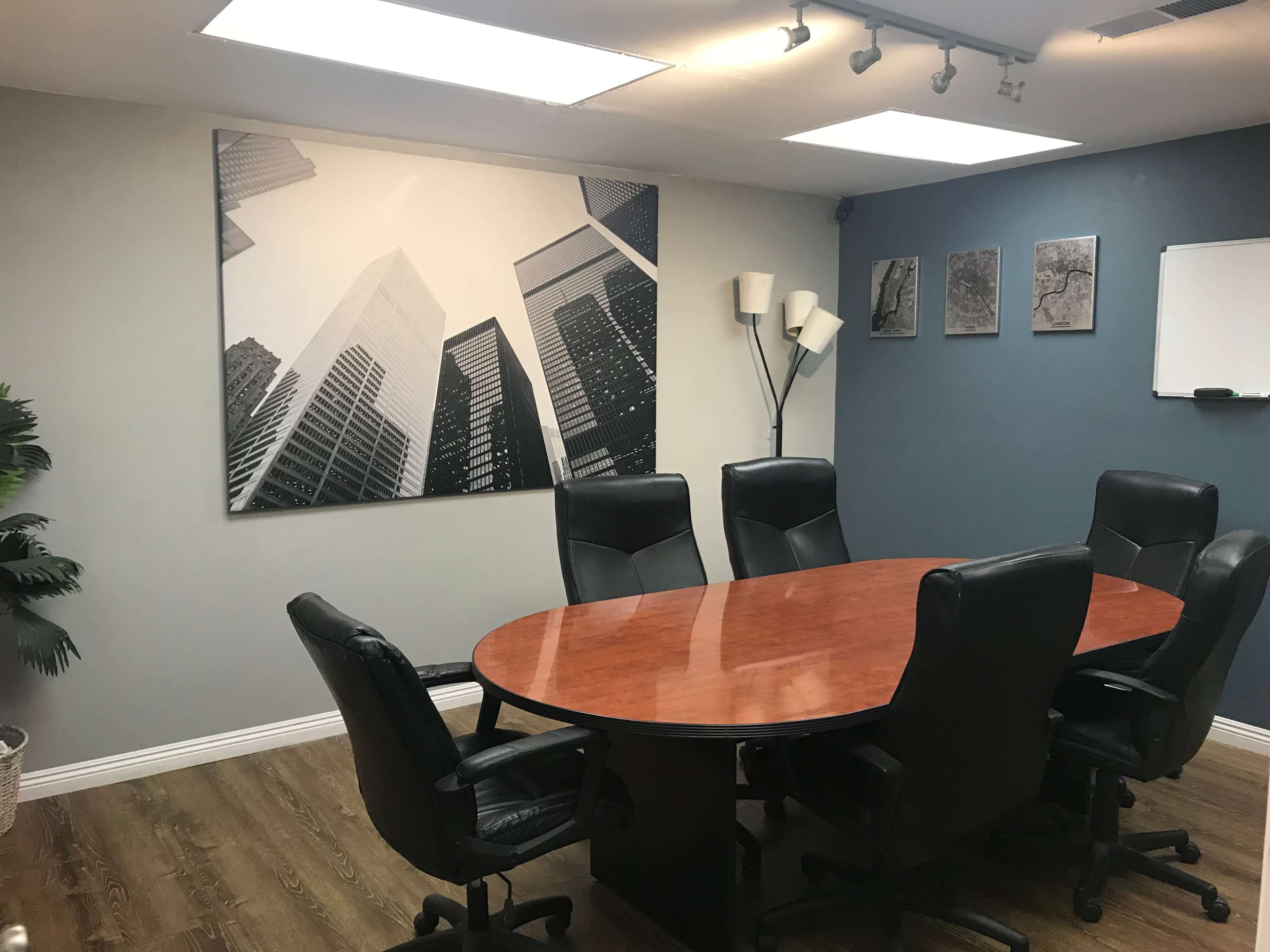 A conference room features a large wooden oval table surrounded by six black leather chairs, with a modern black-and-white cityscape artwork on the wall.