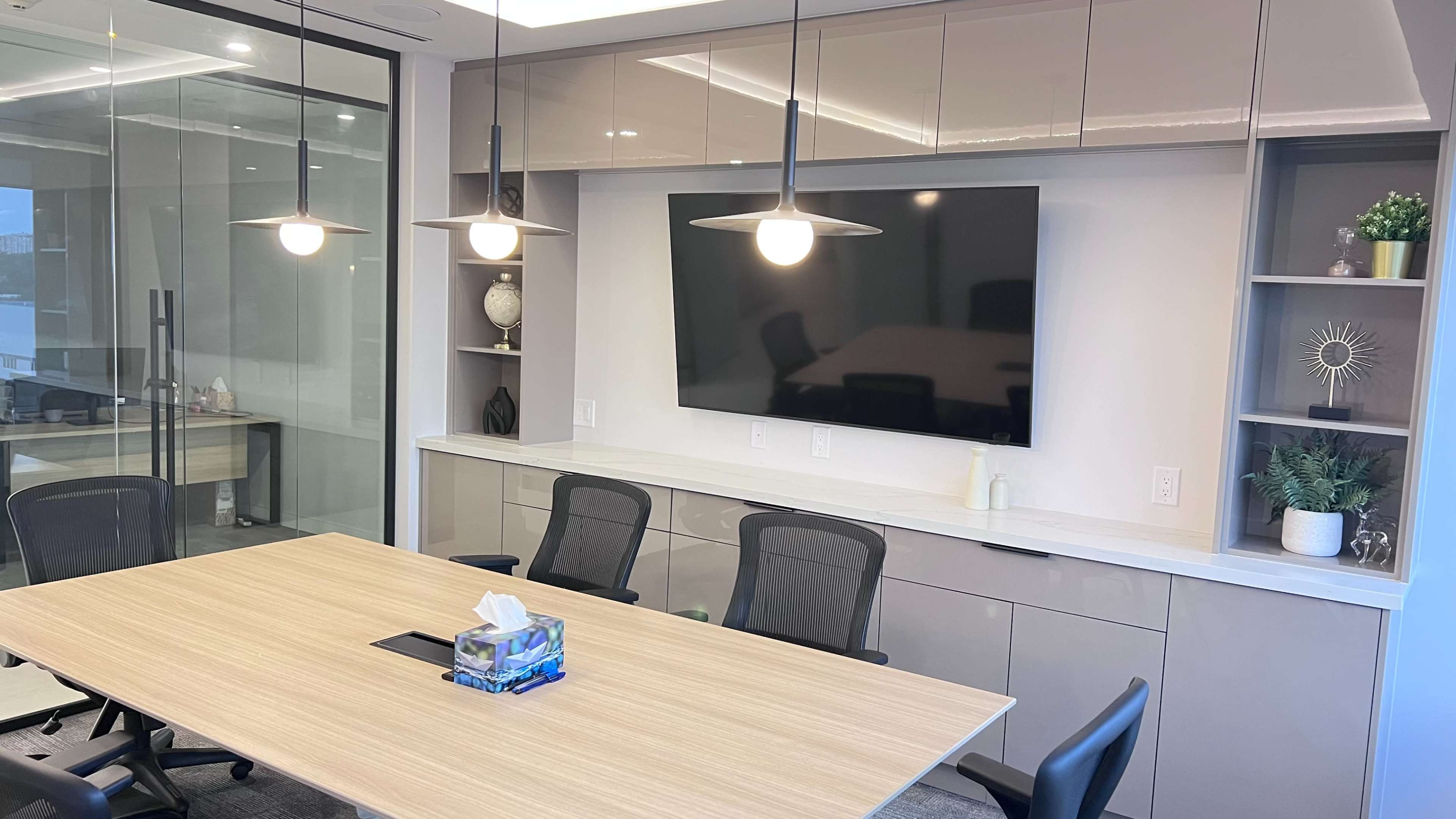 The image shows a modern conference room with a large wooden table, black ergonomic chairs, a wall-mounted TV, and built-in shelving displaying decorative items.