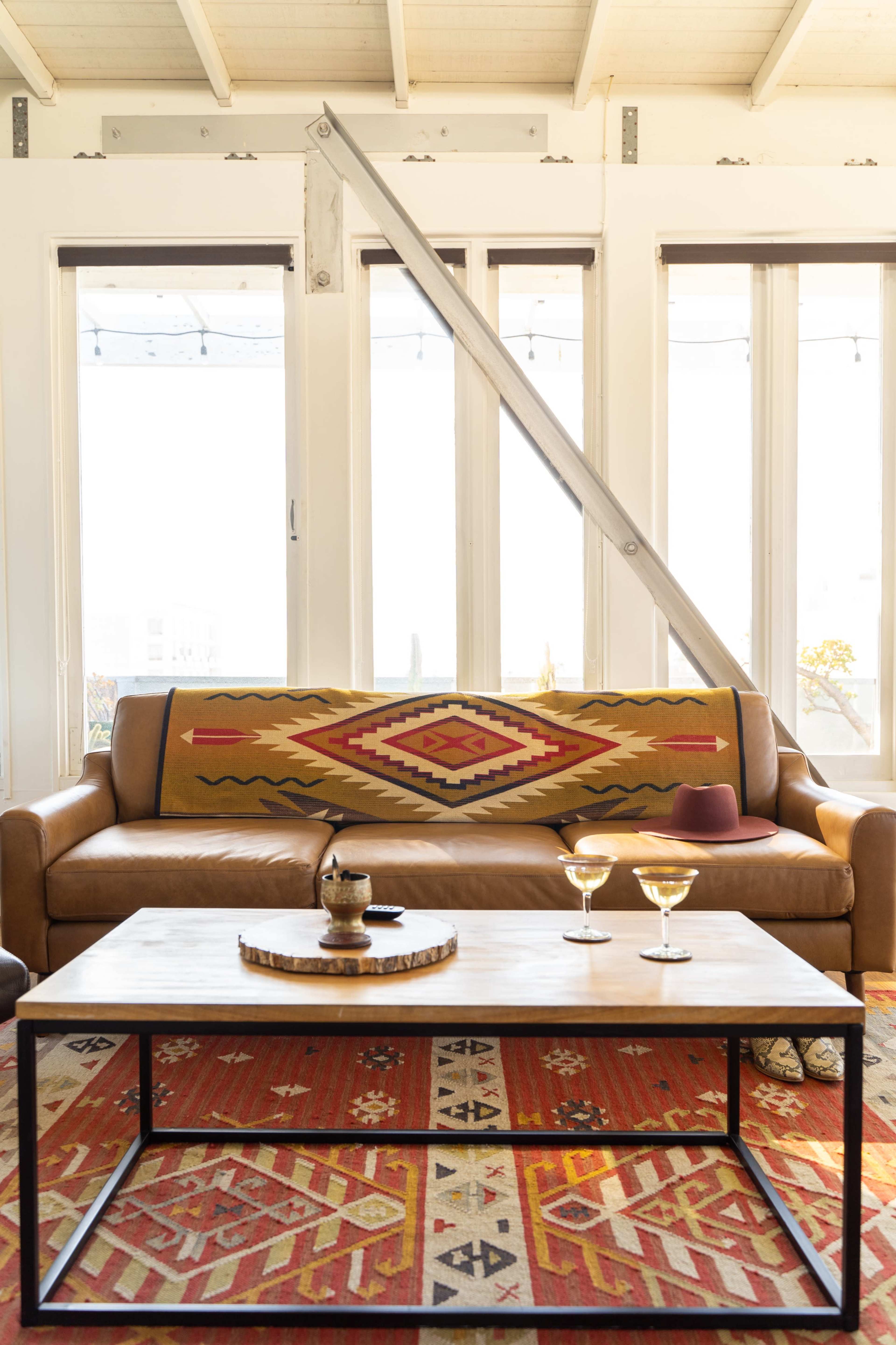 The image shows a modern living room with a brown leather sofa draped with a patterned blanket, a coffee table in front featuring two cocktails, and large windows allowing natural light to fill the space.