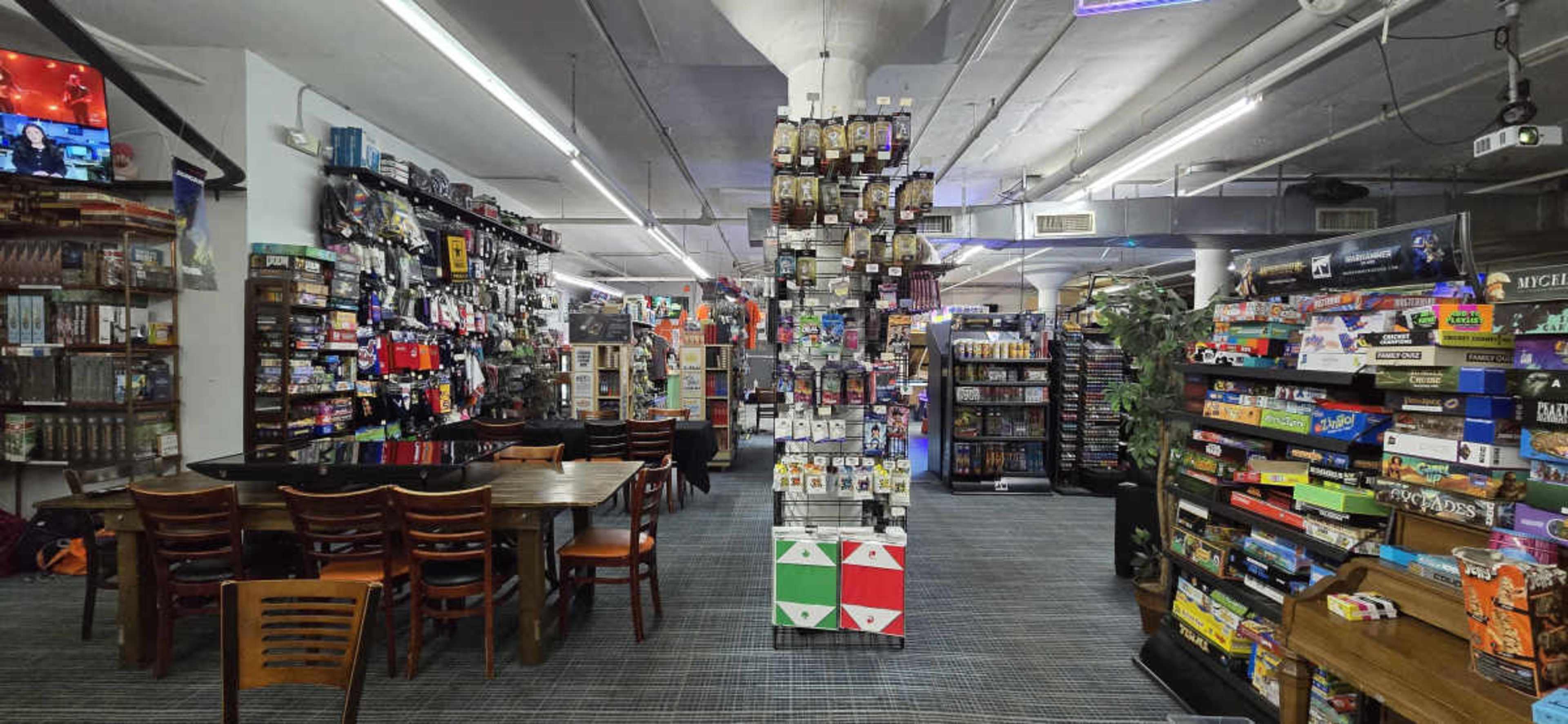 The image shows a well-organized gaming retail space with shelves displaying various board games and gaming accessories, along with a seating area featuring wooden tables and chairs.