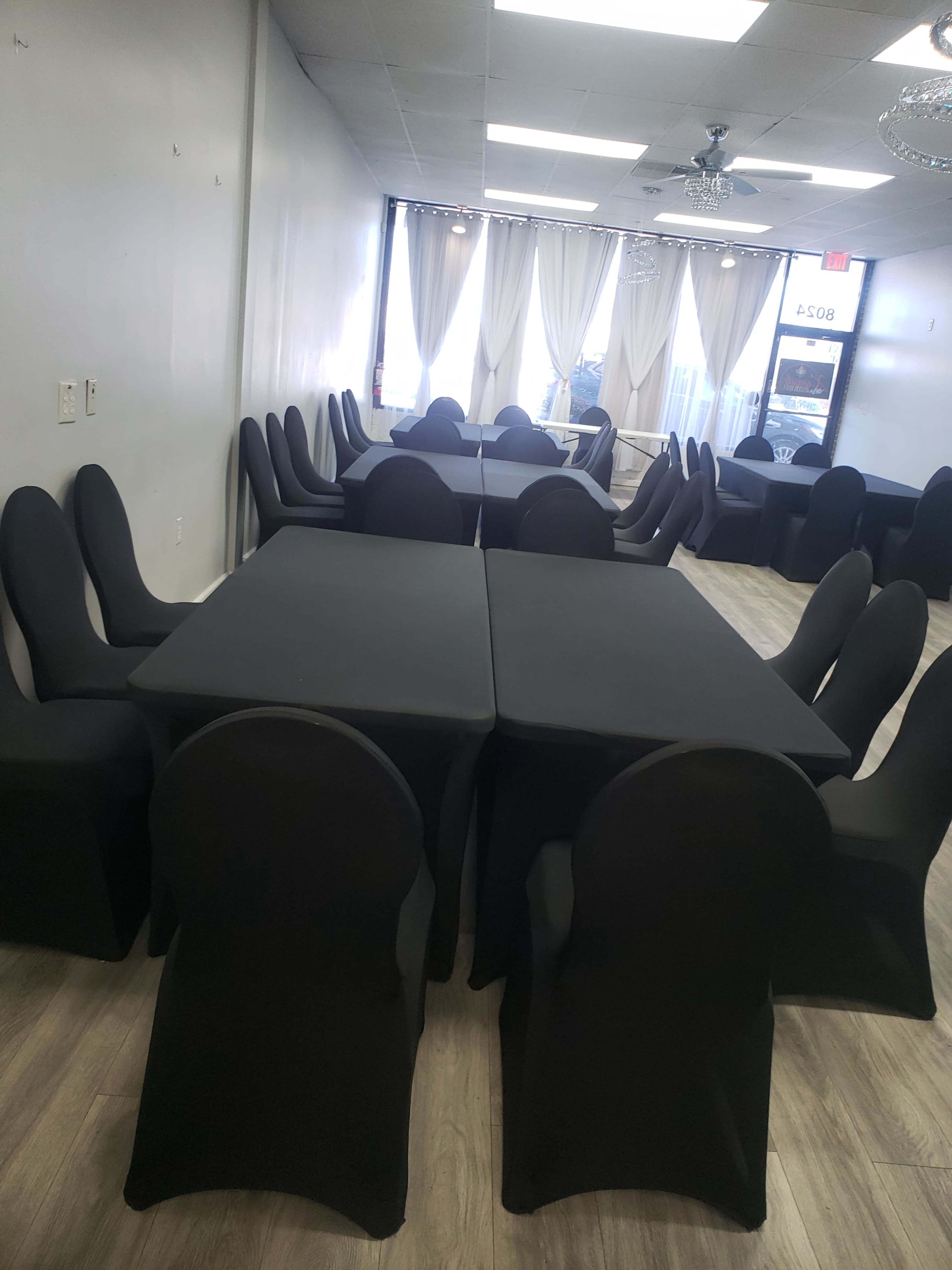 A spacious room with multiple tables and chairs covered in black drapes, arranged for a dining or event setup.