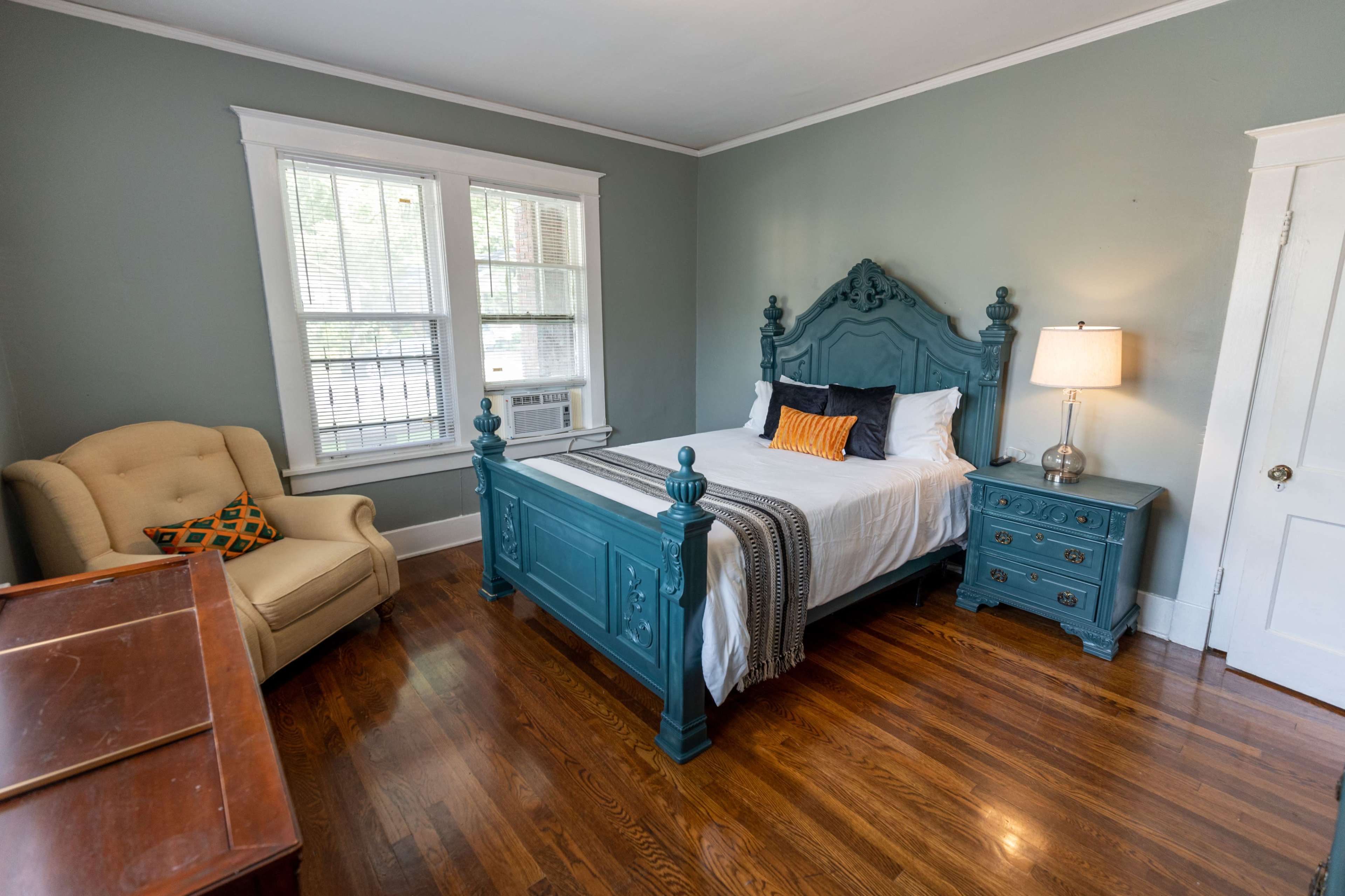 The room features a wooden bed frame with a headboard, an armchair, a nightstand with a lamp, and hardwood flooring.