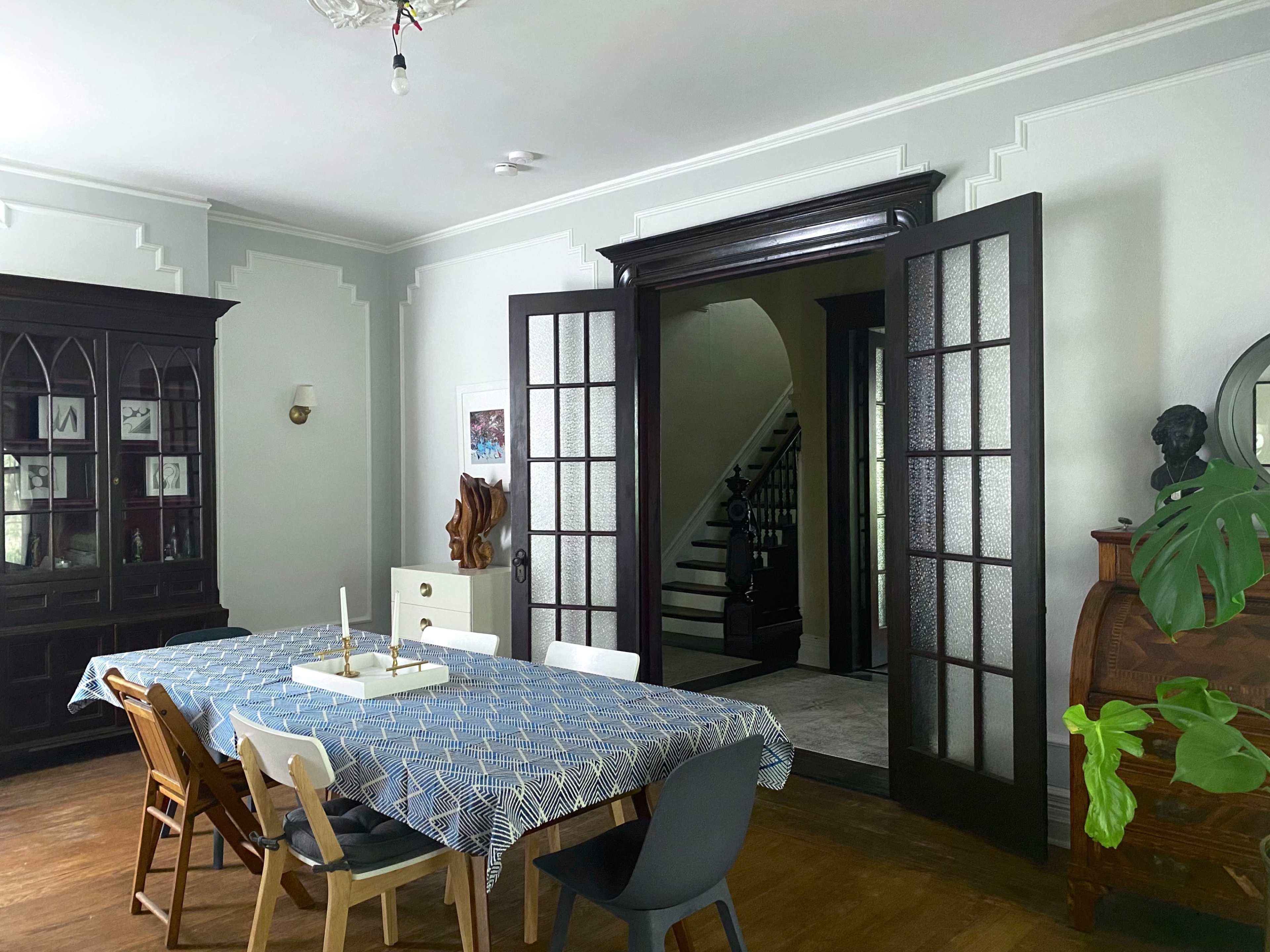 A dining room features a large table with a blue tablecloth, surrounded by various chairs, and an open doorway leading to a staircase.