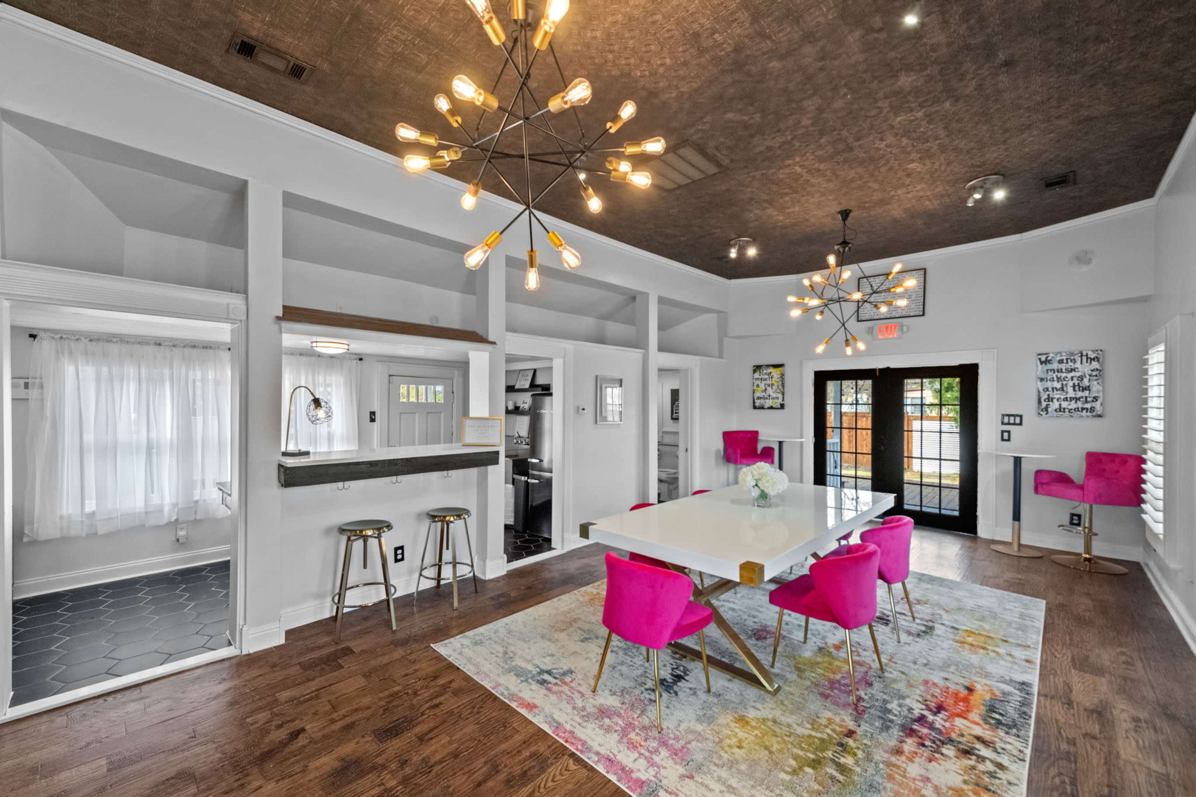 A modern dining area features a large white table surrounded by pink chairs, with decorative light fixtures overhead and large windows allowing natural light to enter.