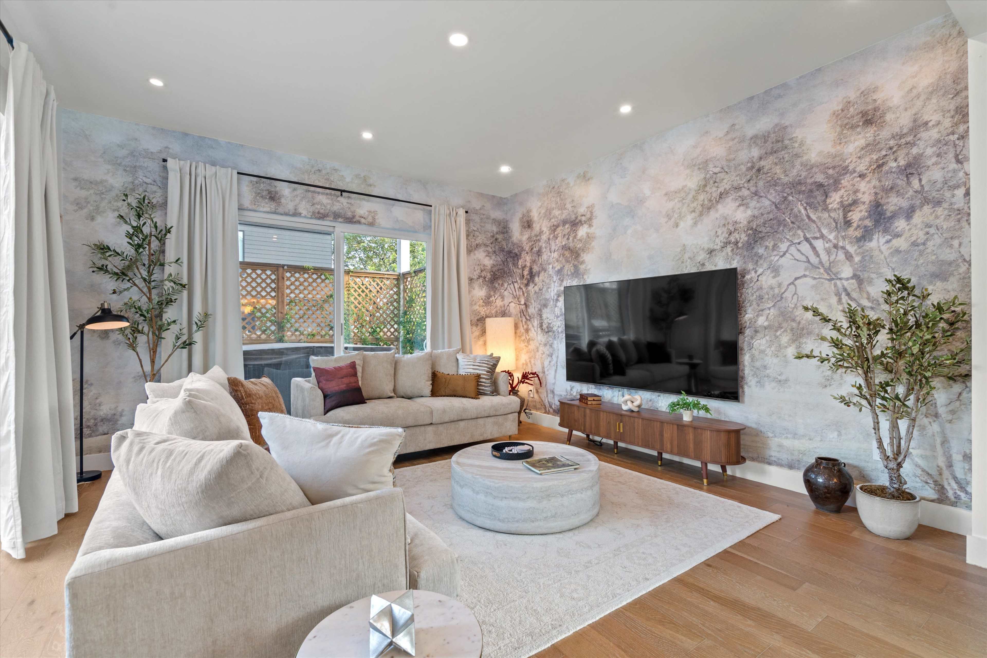 The image shows a modern living room with a large flat-screen TV mounted on the wall, a light-colored sectional sofa, a round coffee table, and decorative plants, all set against a mural depicting a serene nature scene.