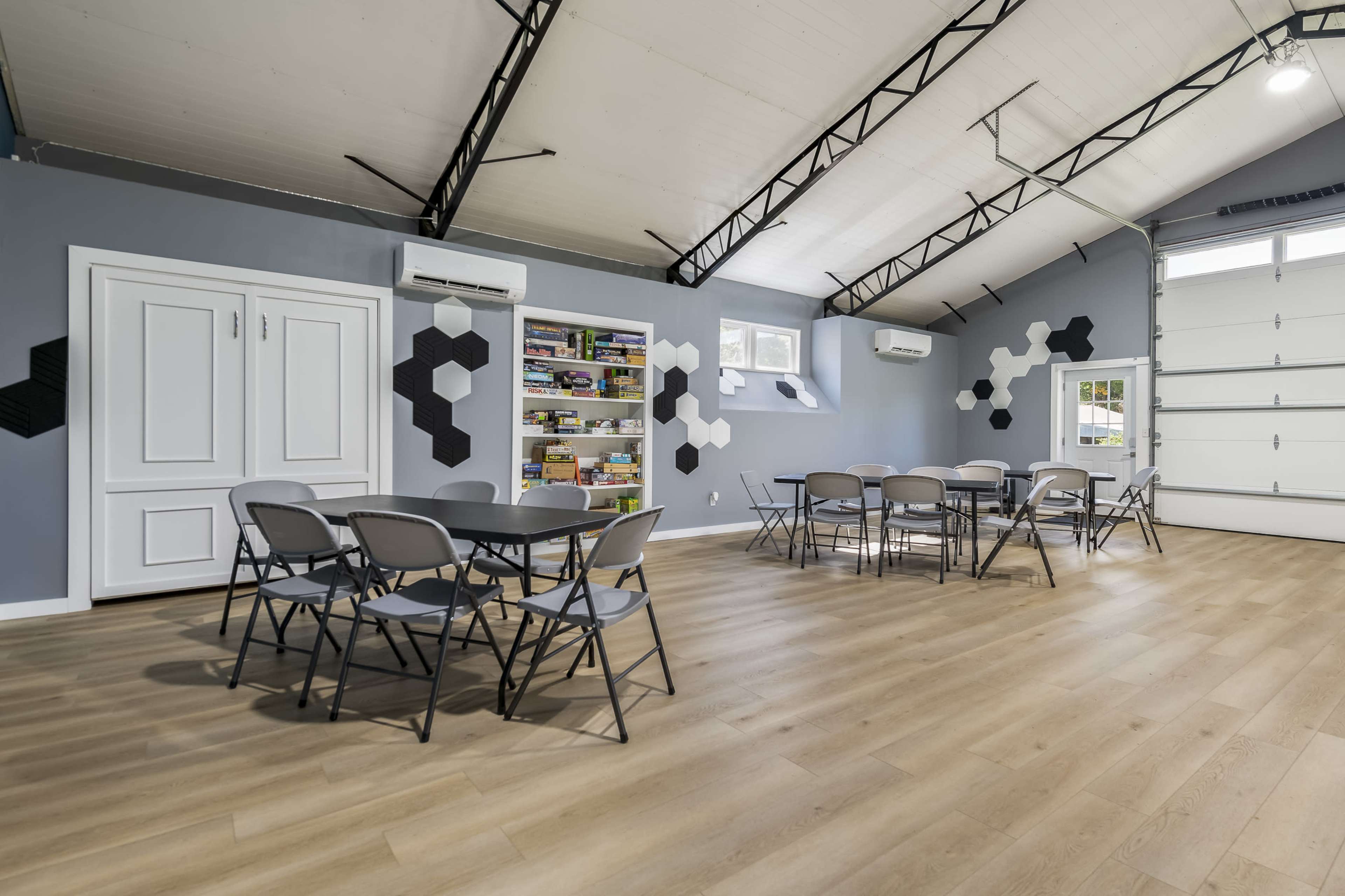 The image shows a spacious room with a high ceiling, featuring gray walls, a shelving unit filled with board games, and several round tables with chairs arranged for seating.