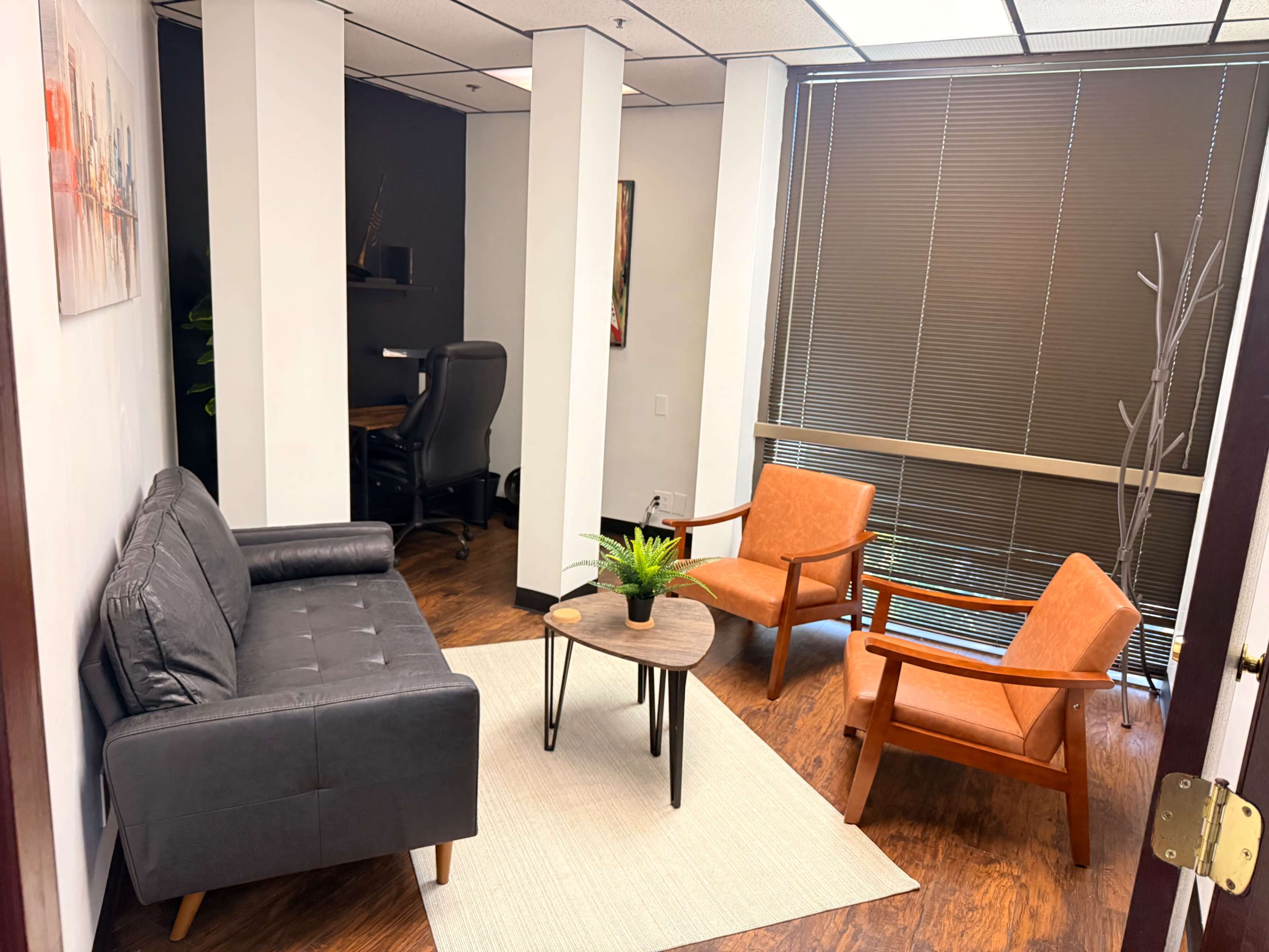 The image depicts a small office waiting area with a gray couch, two orange armchairs, and a round table on a light rug.