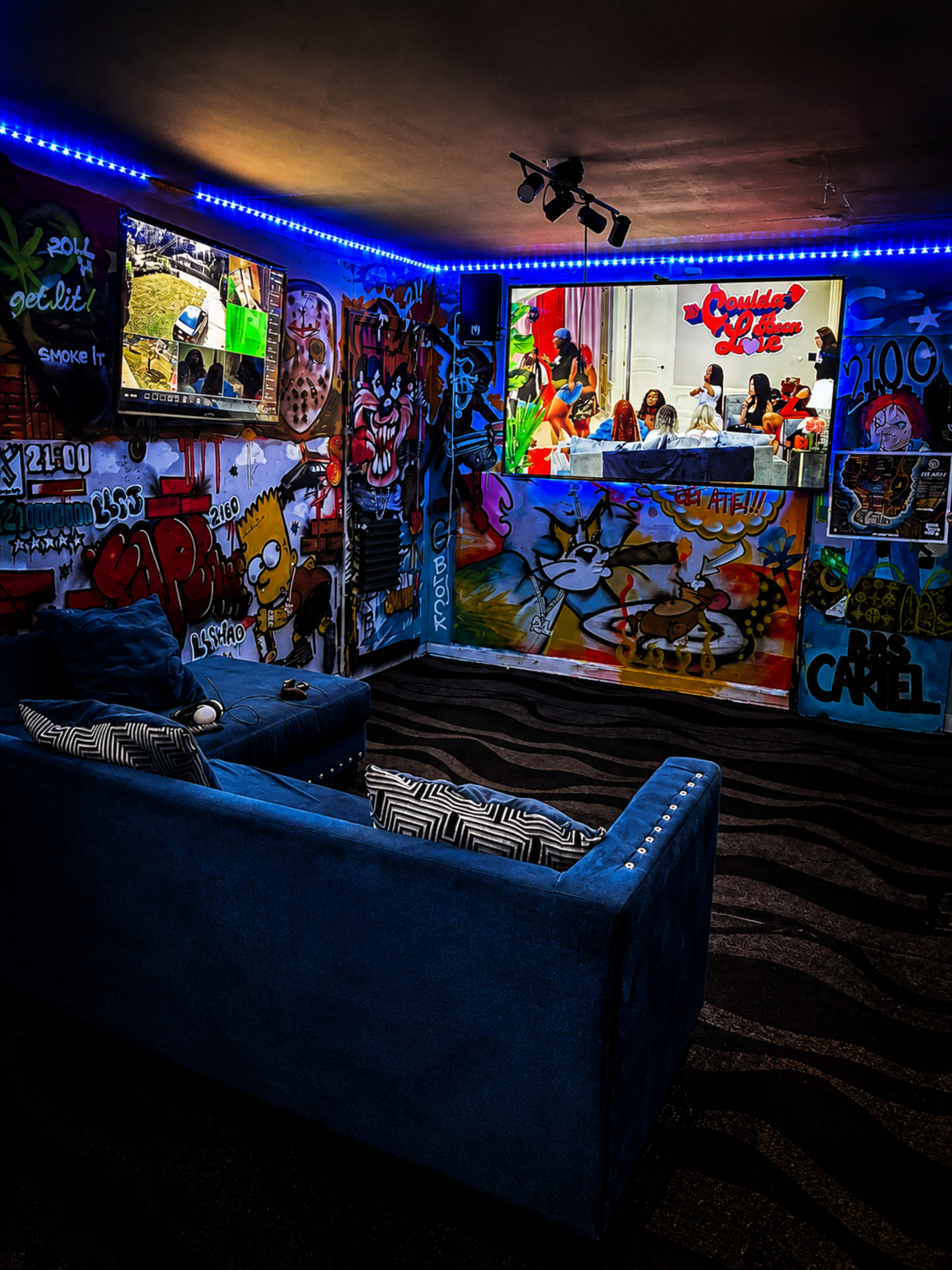 The image shows a colorful graffiti-decorated room featuring a large screen displaying a group of people, with blue sofas placed in front on a striped carpet.