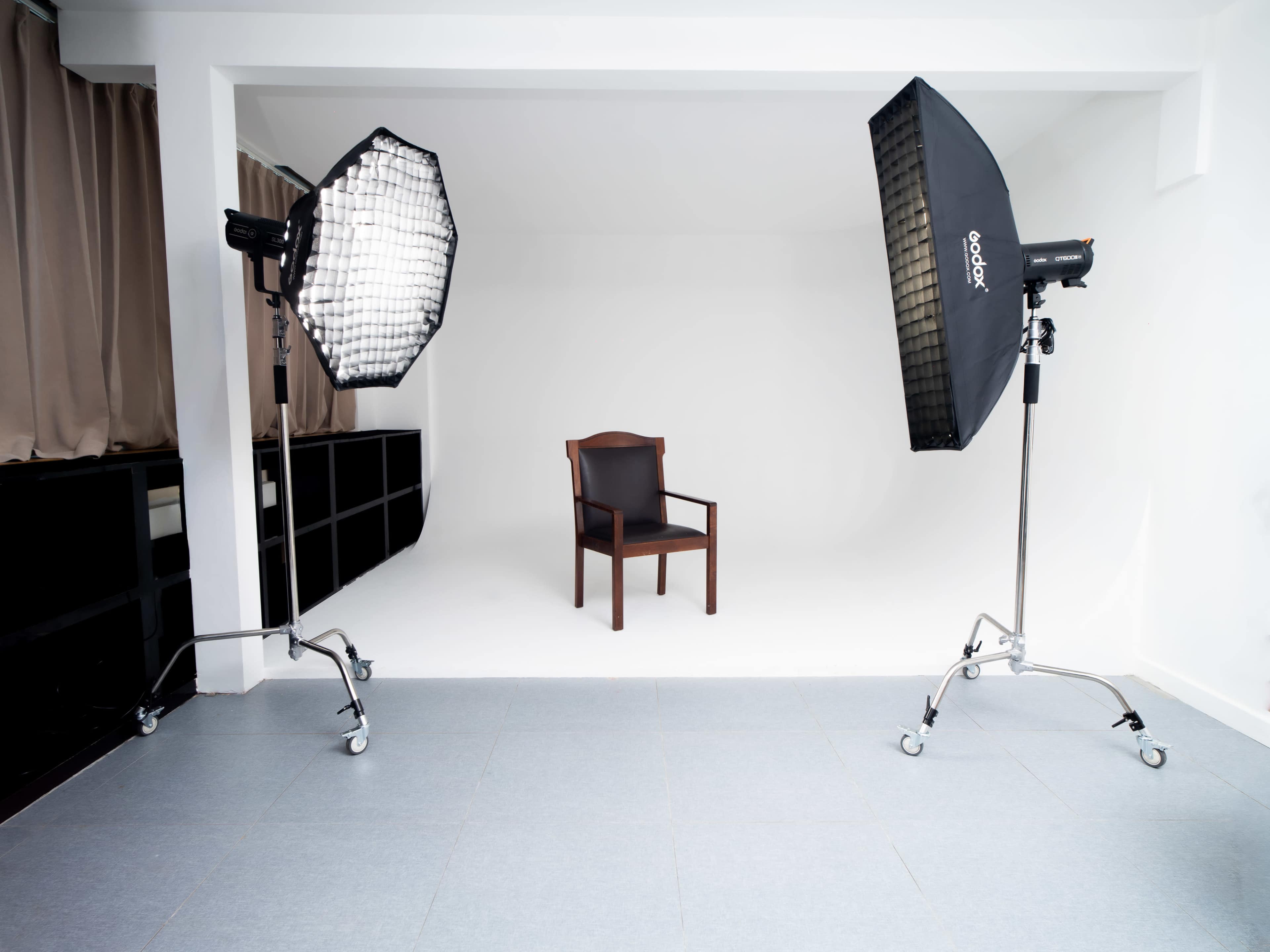 The image shows a minimalist photo studio featuring a single brown chair positioned in front of a plain white background, with two softbox lights on either side.