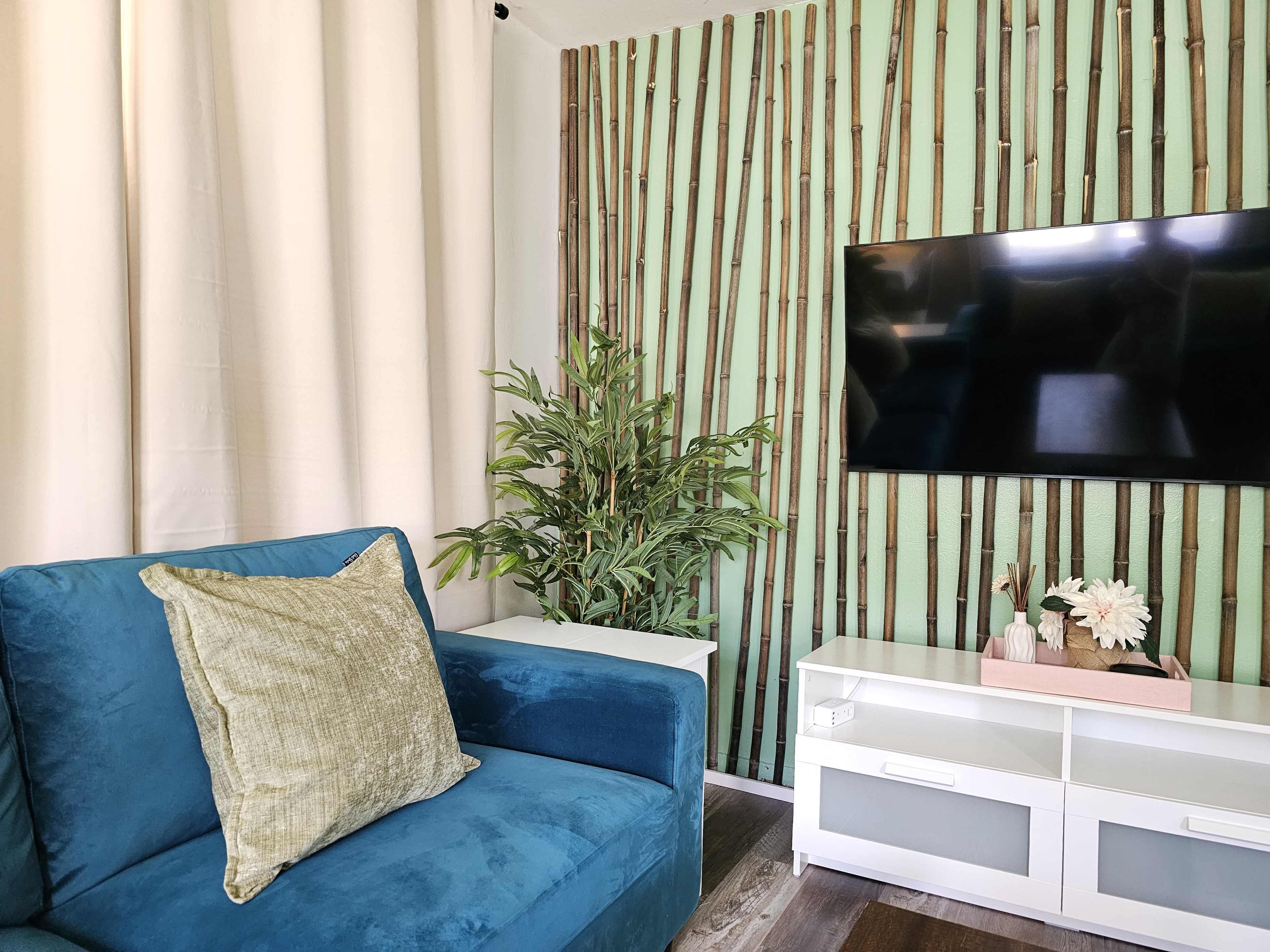 A living room features a blue sofa with a green pillow, a tall indoor plant, a flat-screen TV on a light-colored media console, and a wall decorated with vertical bamboo-like accents.