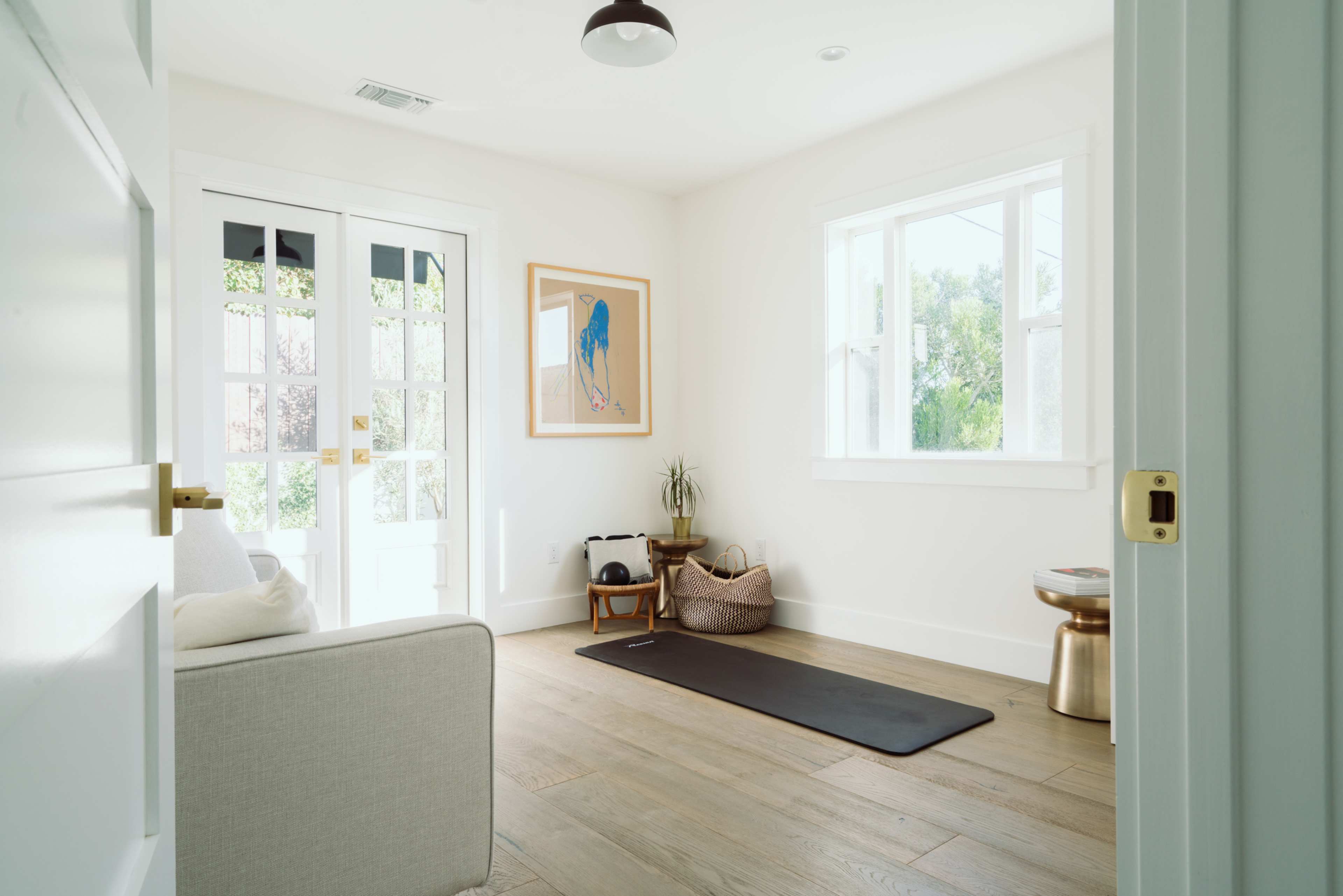 A bright, minimalistic room features a yoga mat on the wooden floor, a small plant, and light streaming through large windows alongside a set of French doors.