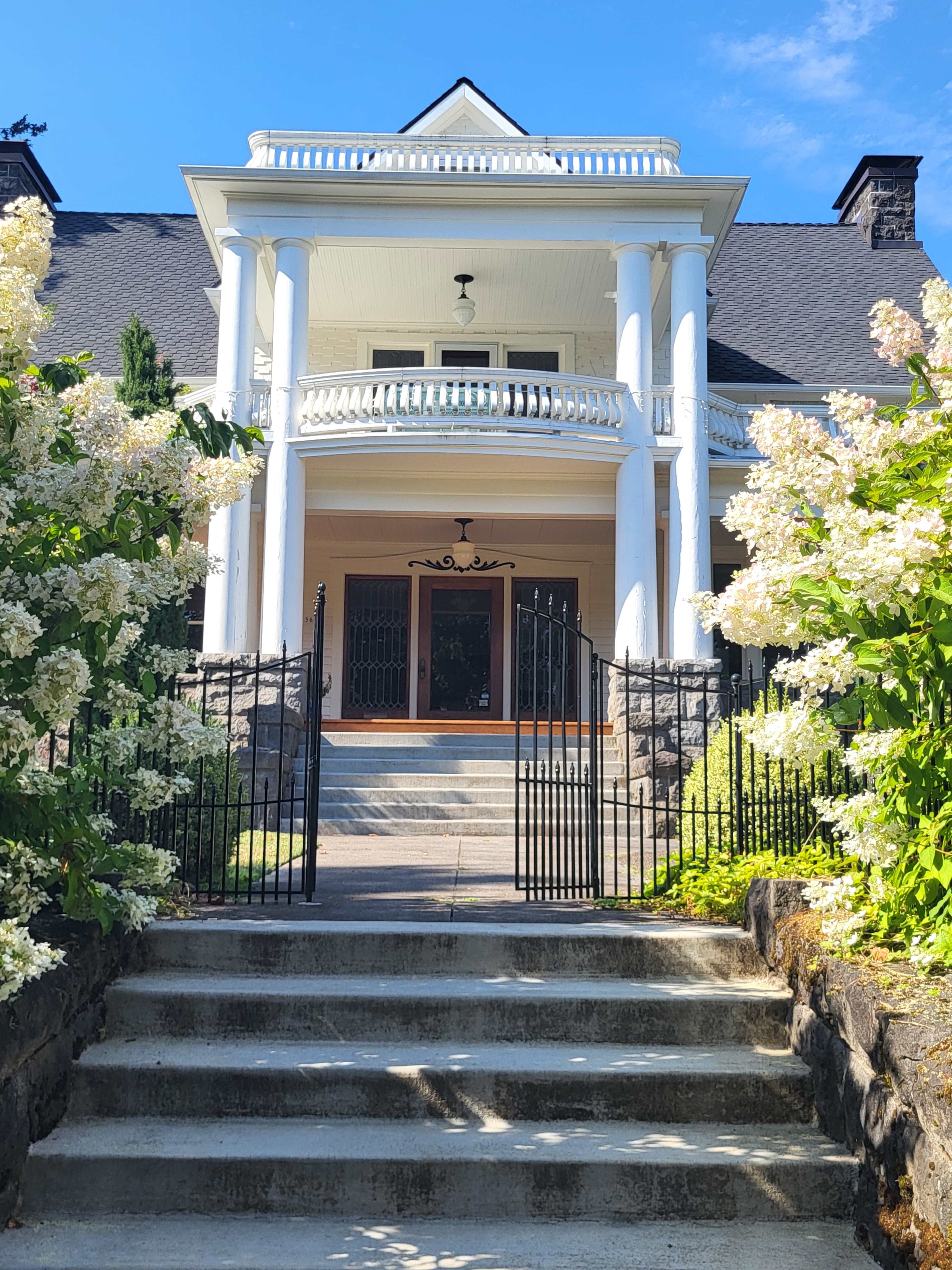 A grand house with white columns stands at the top of a stone staircase, surrounded by blooming hydrangeas and framed by a decorative wrought-iron gate.