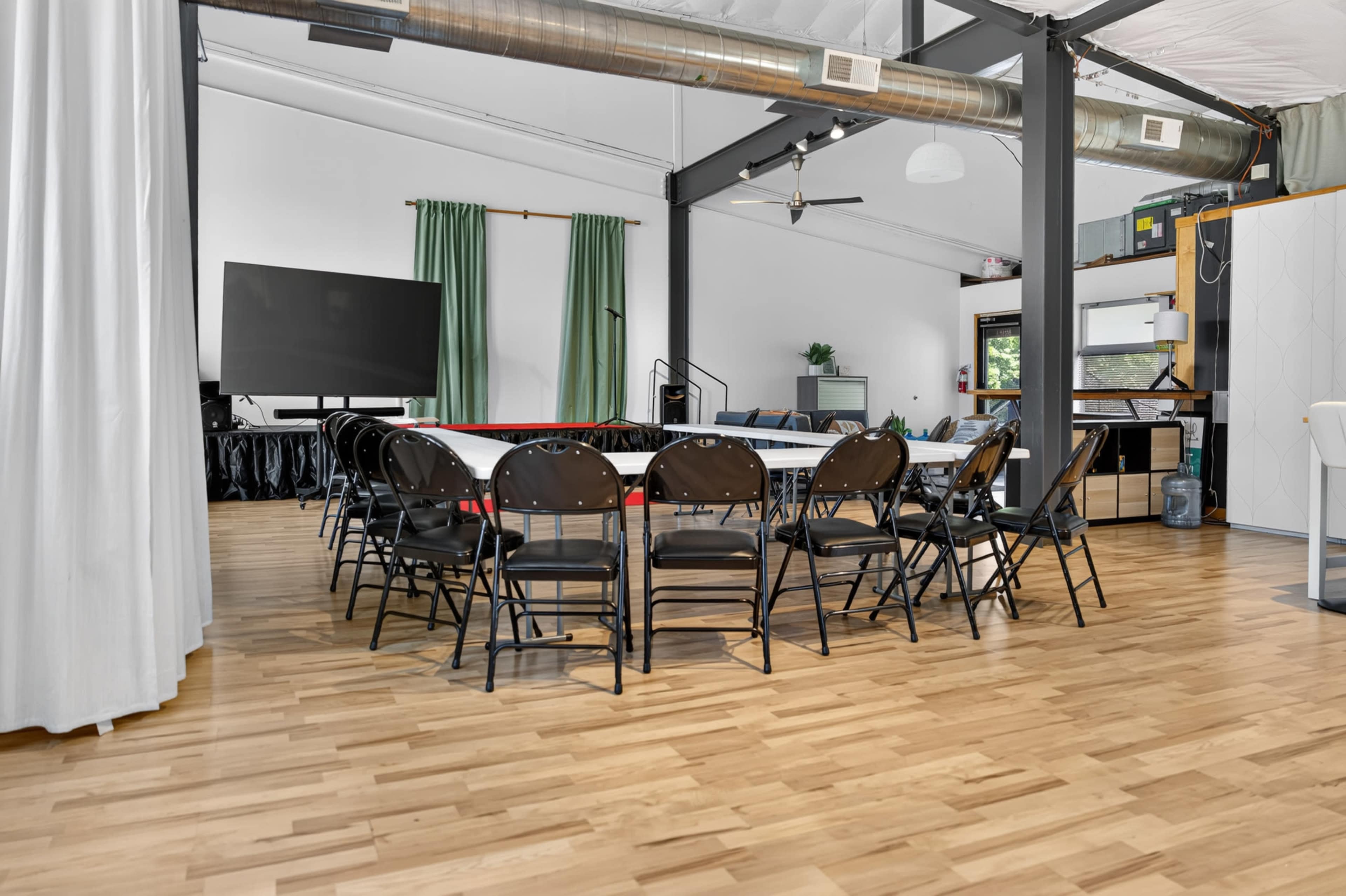 A conference room features a large circular table surrounded by black folding chairs, with a large screen in the background and a spacious layout.