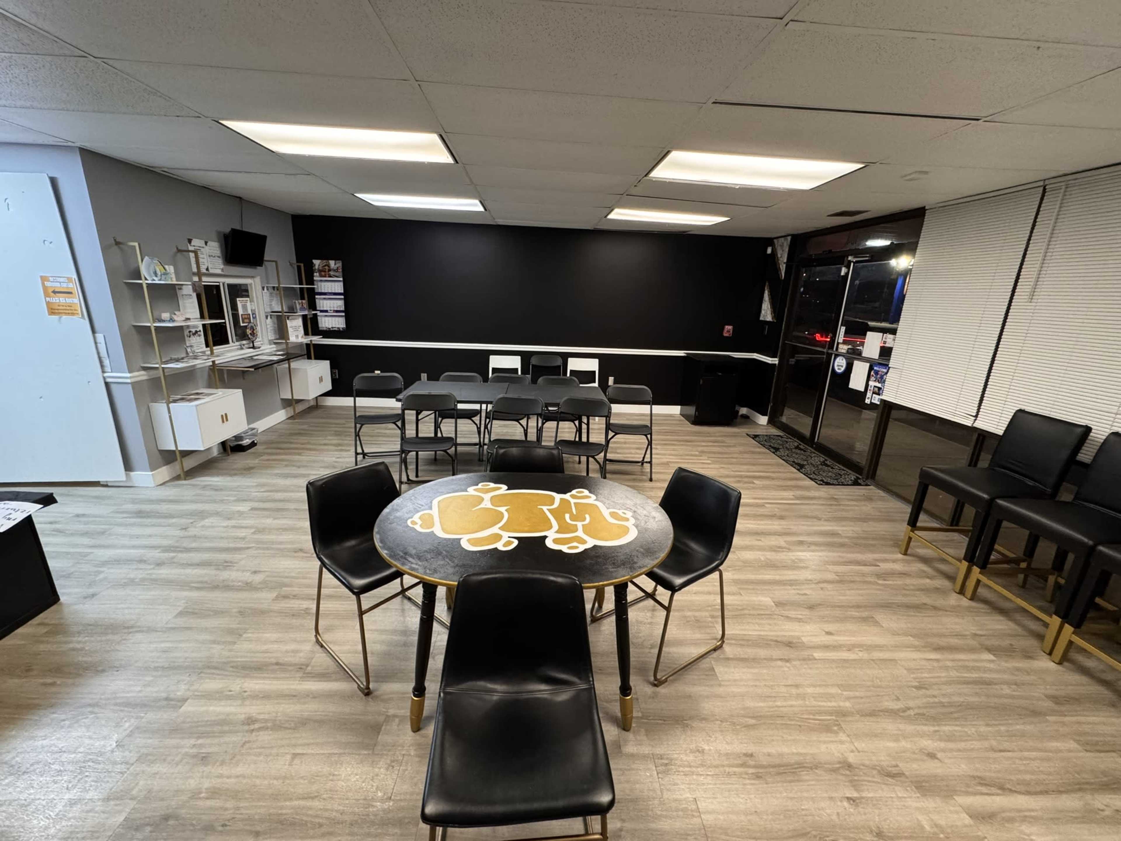The image shows a spacious room with a circular table in the center, surrounded by black chairs, and a few additional tables arranged along the walls.