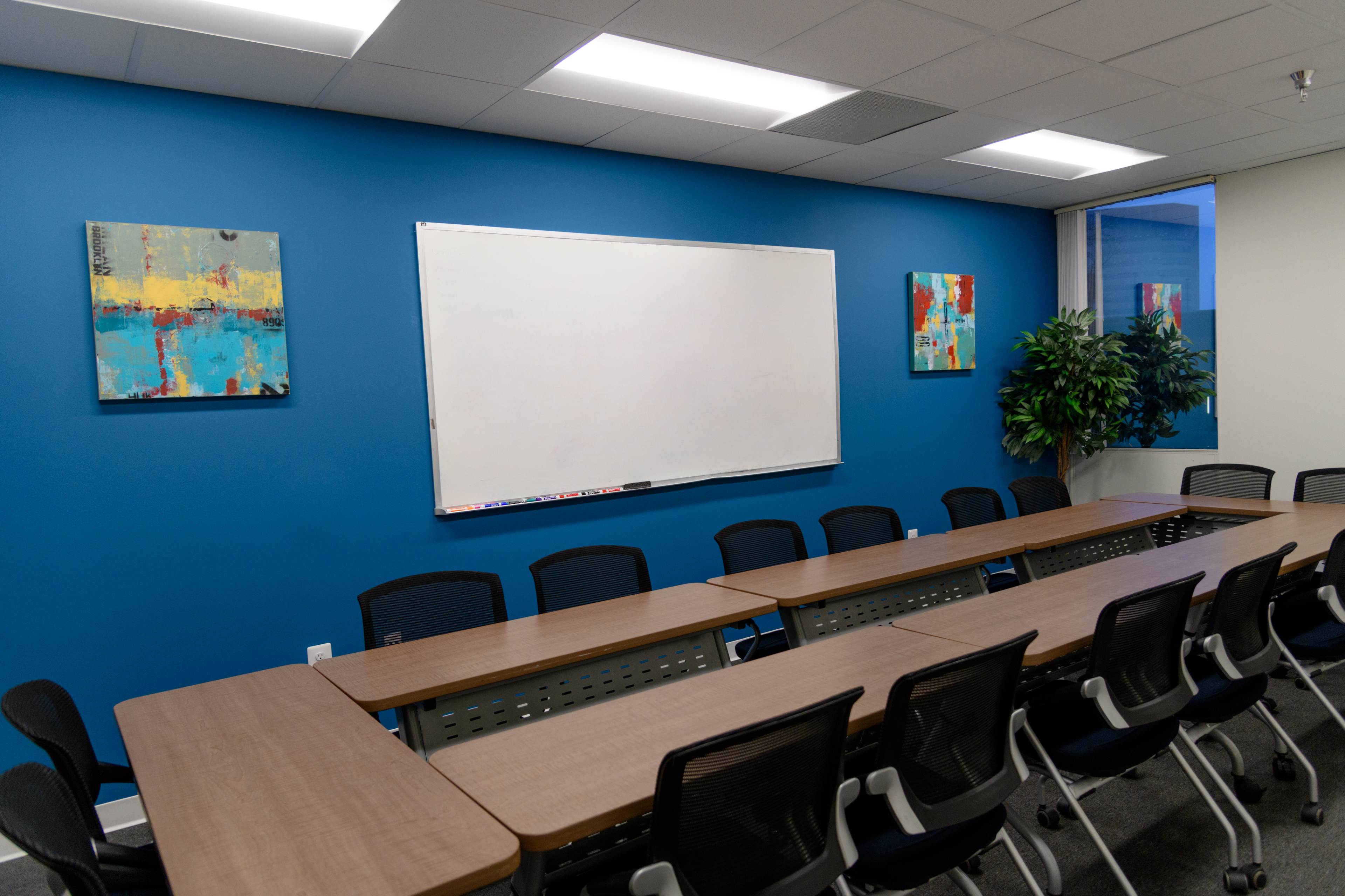 A modern conference room features a long table with several ergonomic chairs, a whiteboard on a blue wall, and two abstract paintings on either side.